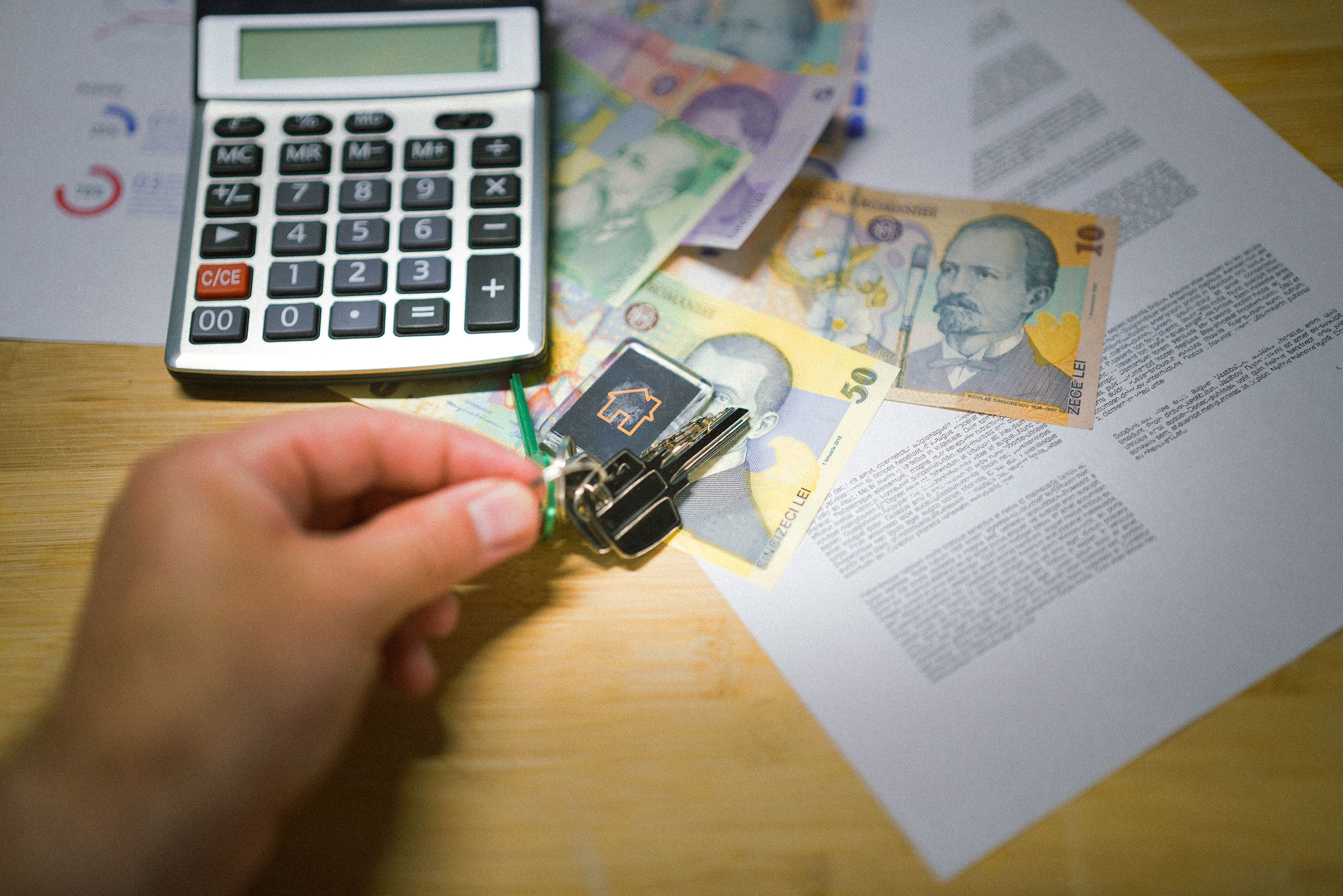 A hand holds keys next to Romanian currency, a calculator, and documents, symbolizing real estate transactions.