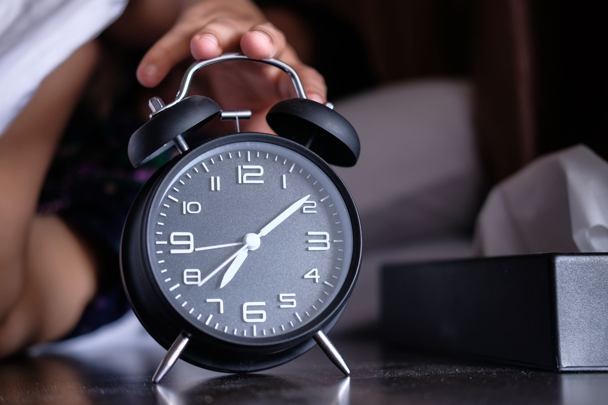 A hand reaches out to turn off an analogue alarm clock in the early morning.