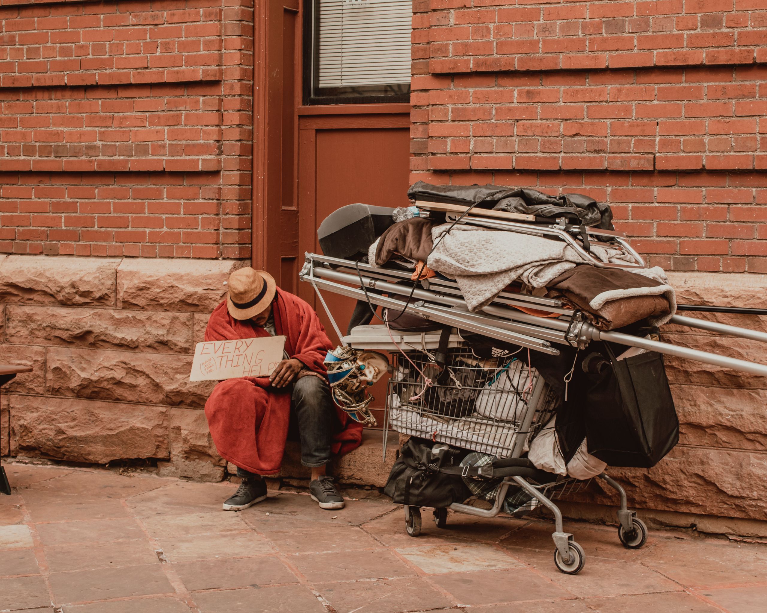 A homeless man sits by a brick wall with a shopping cart, holding a sign.