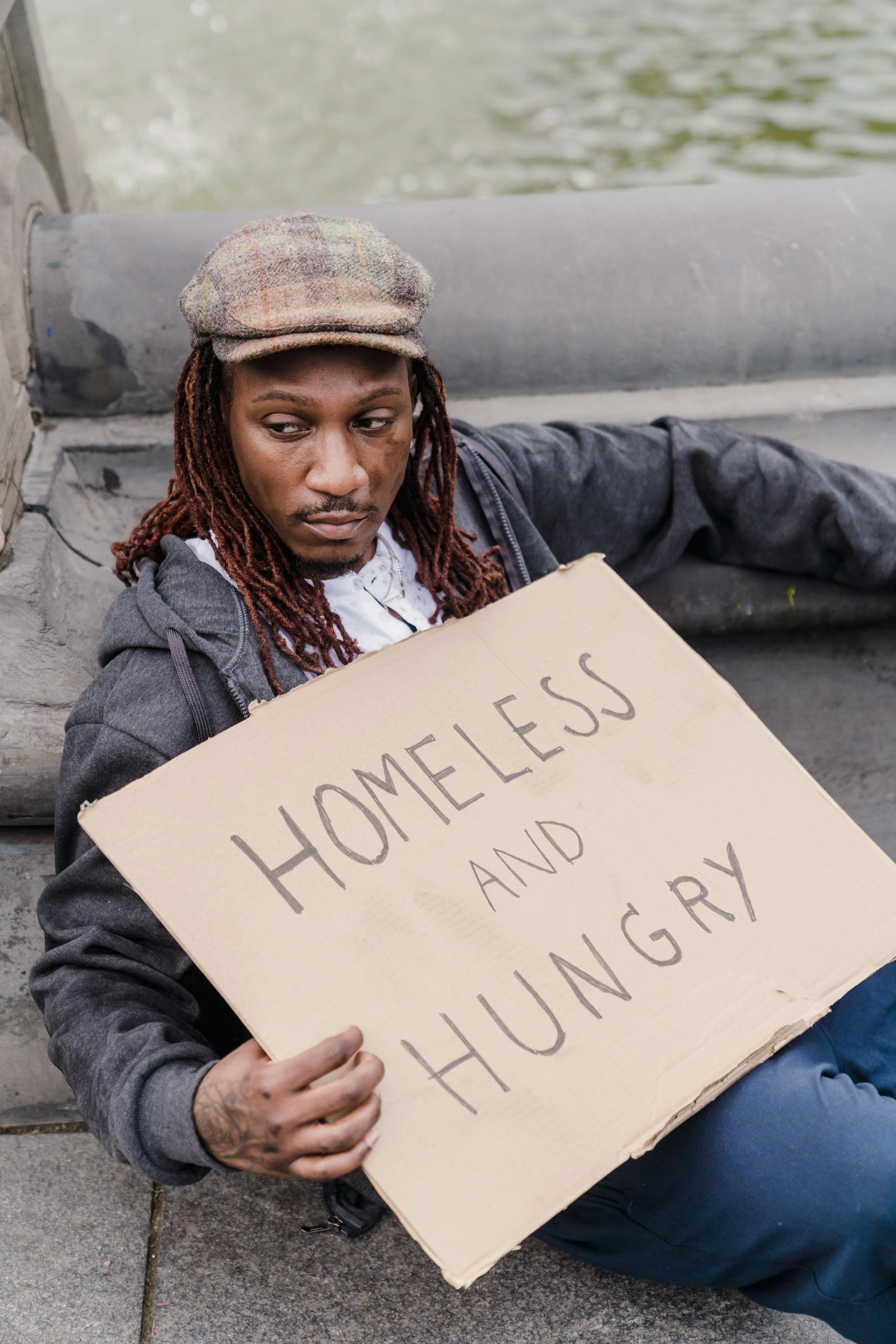 A homeless man sits with a sign reading 'Homeless and Hungry' on the street.
