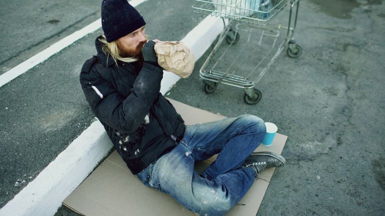 A homeless person seated on a cardboard, drinking from a paper bag outdoors.