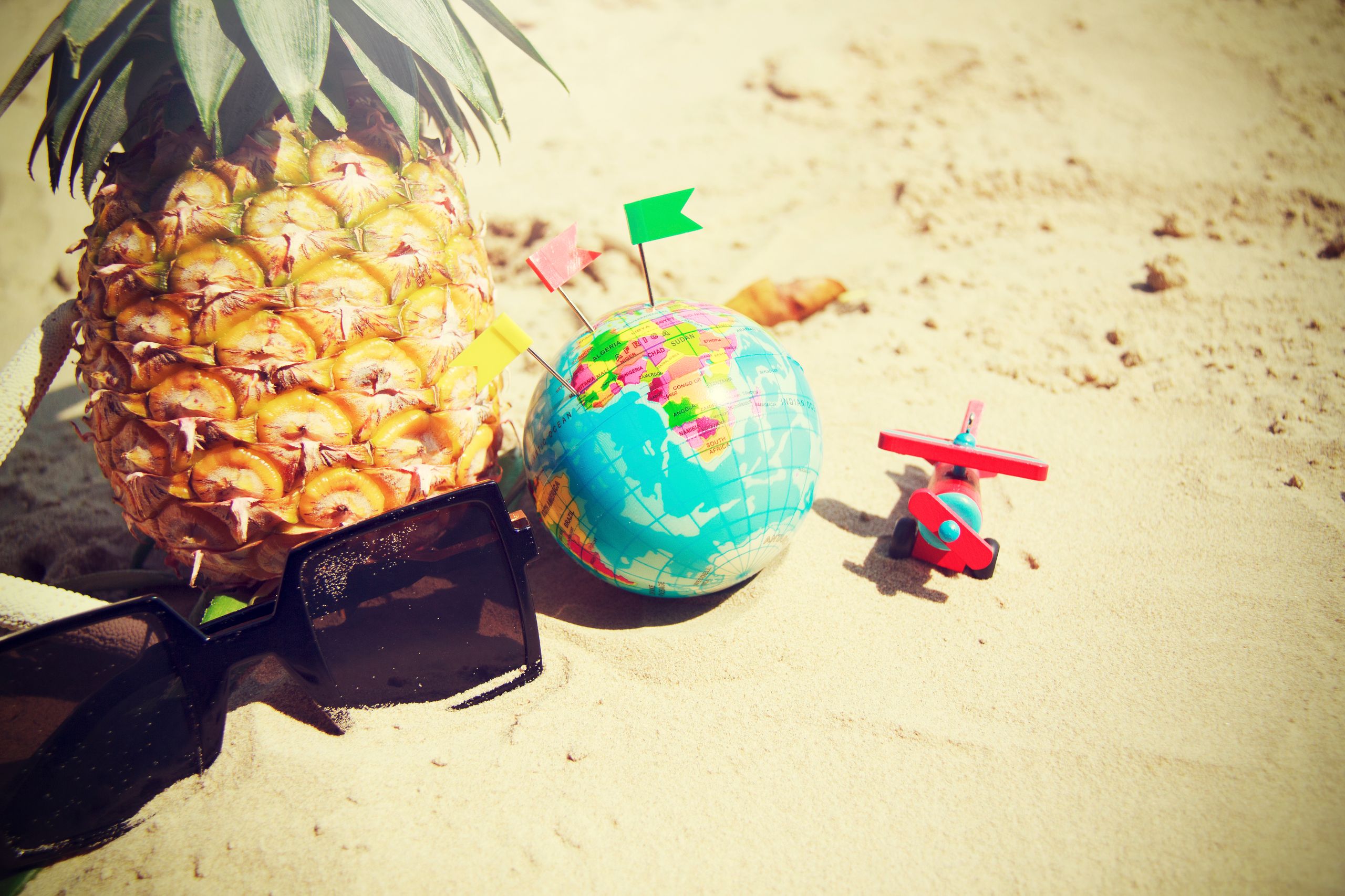 A lively summer composition with a pineapple, globe, and toy plane on a sandy beach.