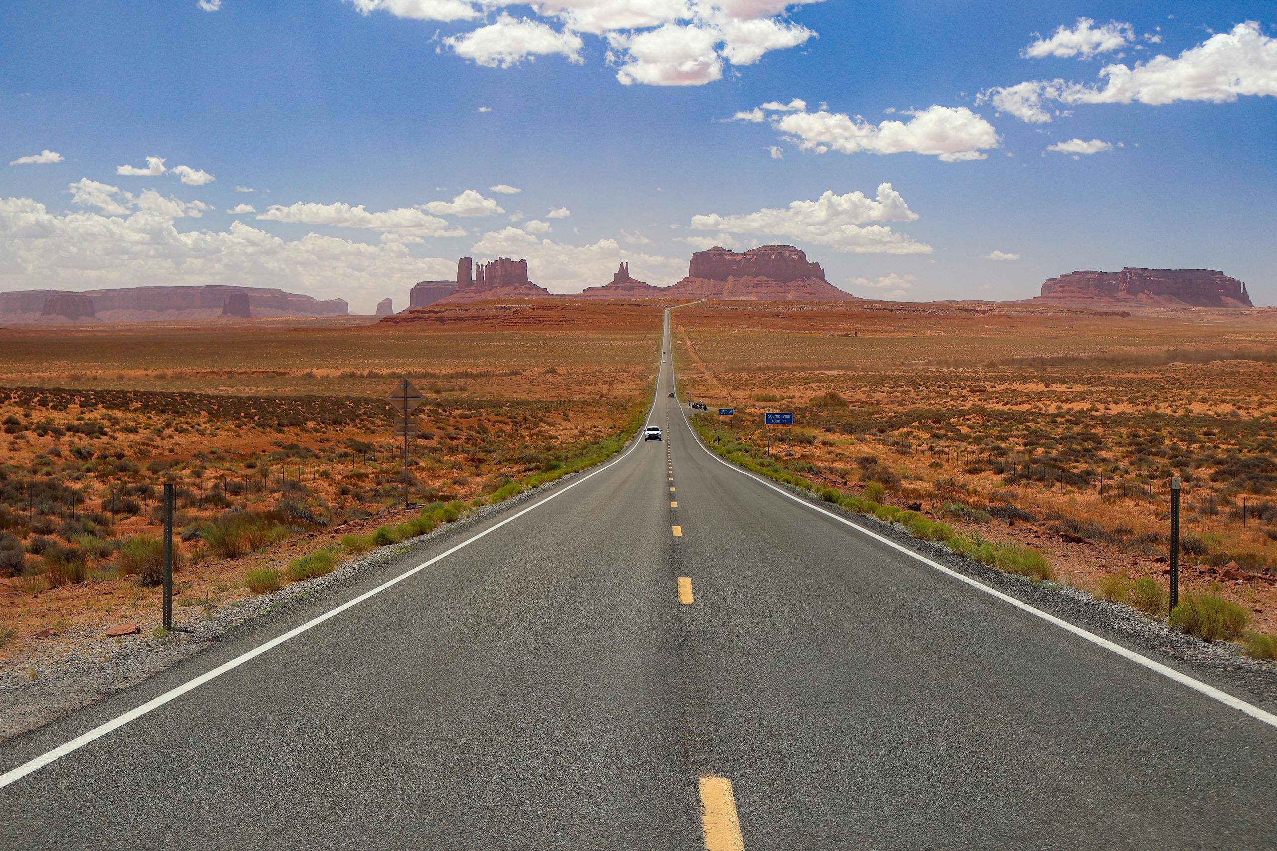 A long, empty highway stretches through Monument Valley under a bright blue sky.