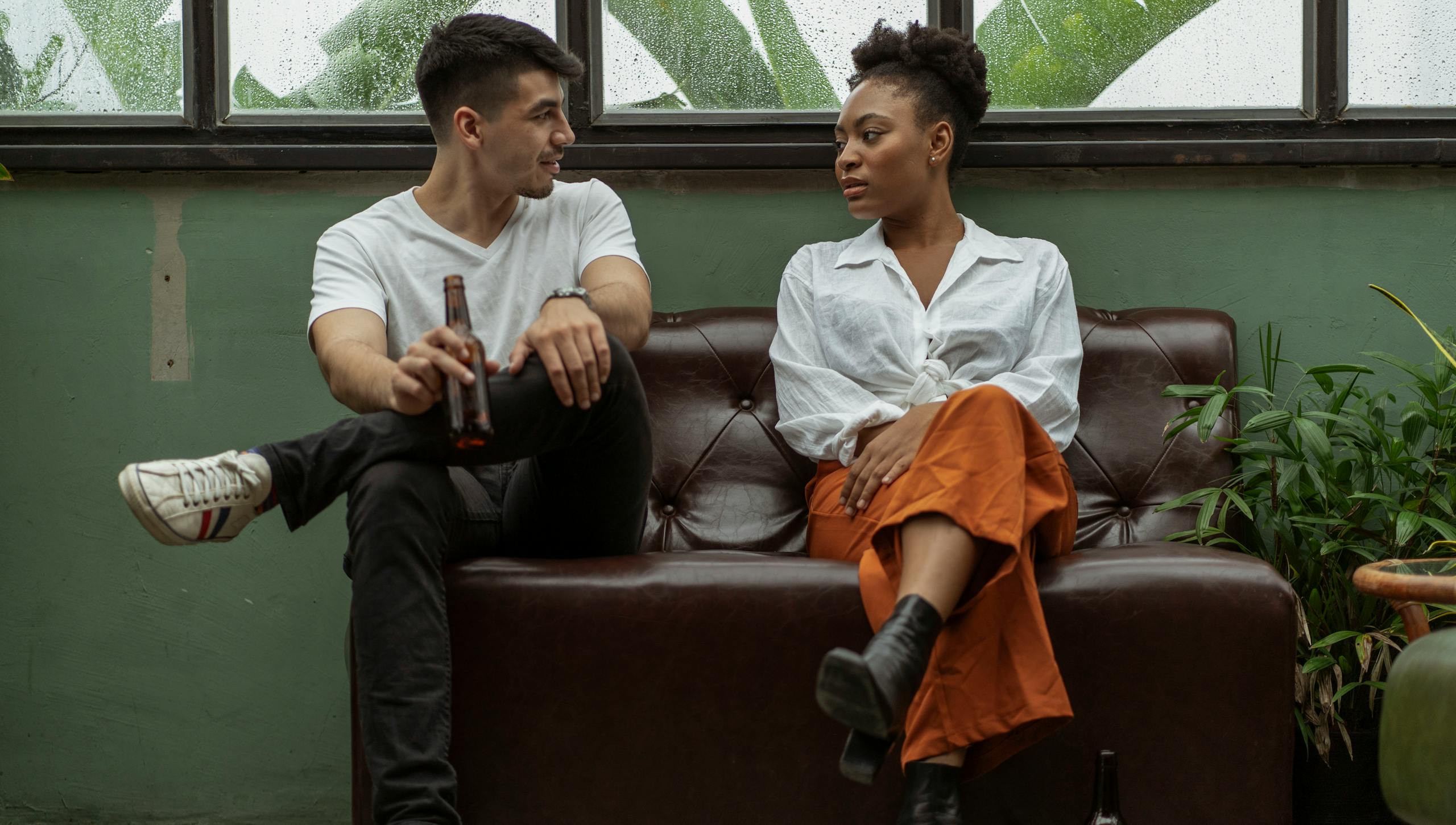 A man and woman engage in a casual conversation while sitting on a sofa indoors, holding beer bottles.