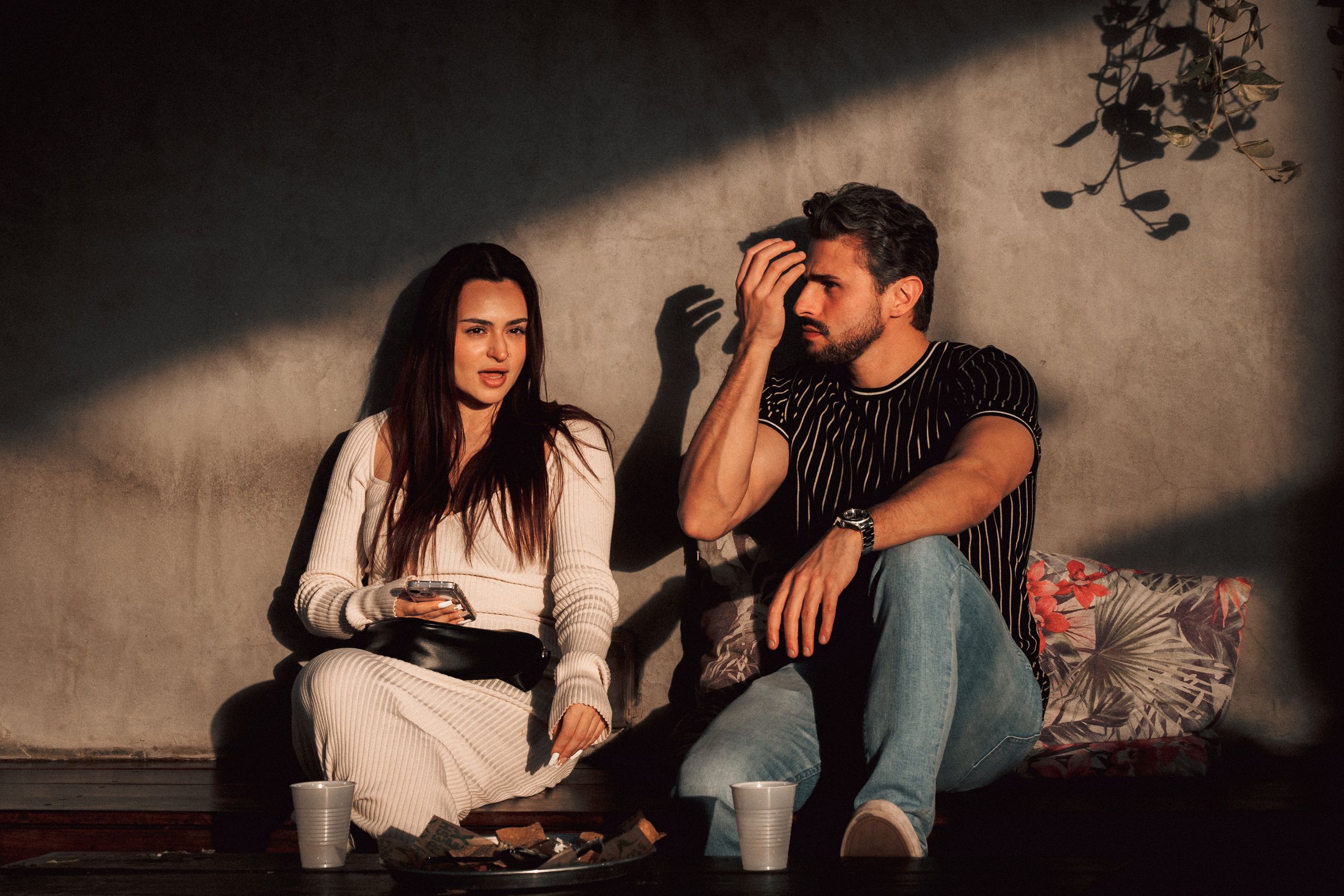 A man and woman sitting outdoors, engaged in casual conversation under natural light shadows.