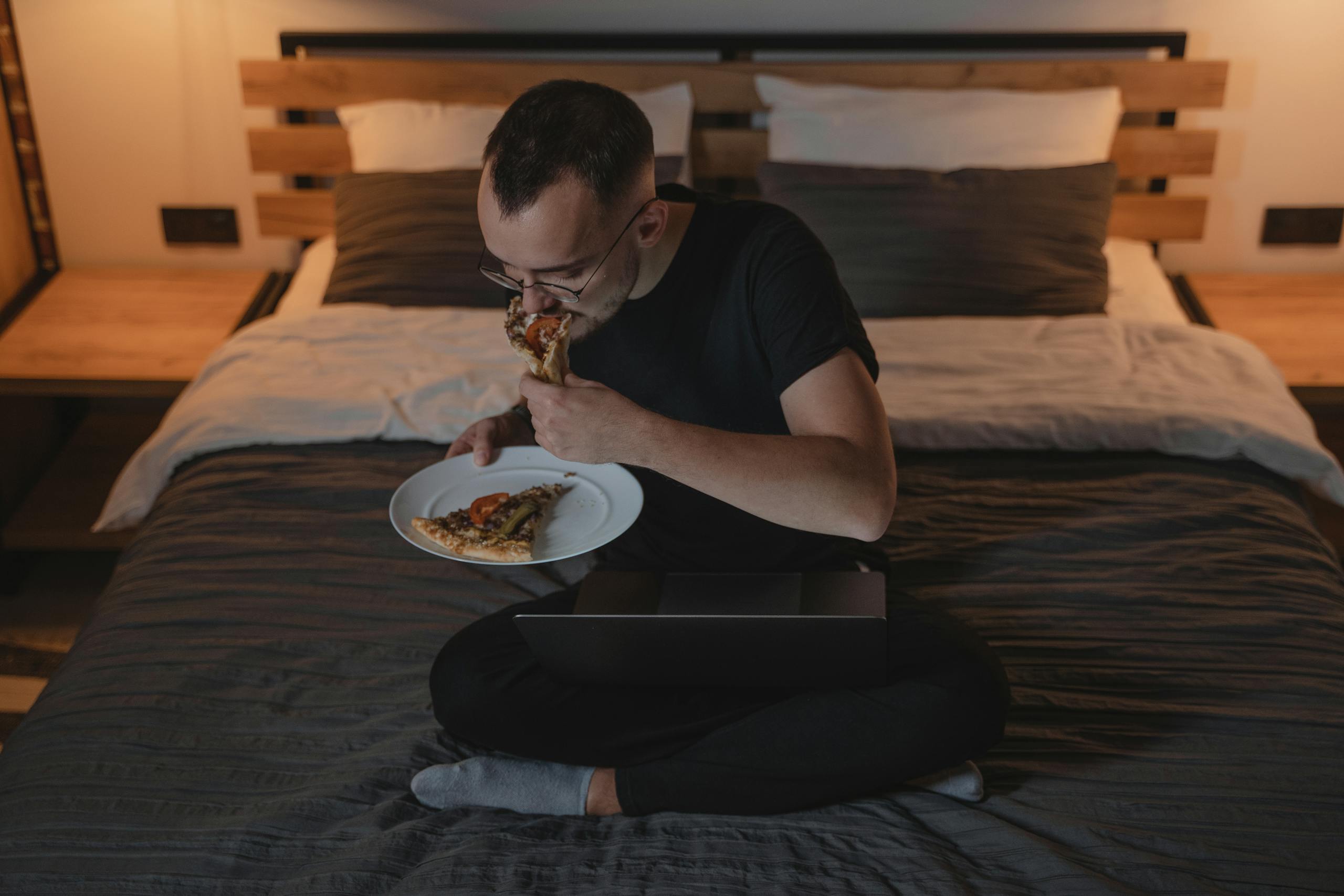 A man enjoying a pizza while working on a laptop in a cozy bedroom setting.
