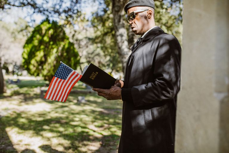 A man in a black coat holding a Bible and US flag during a memorial service in a park.