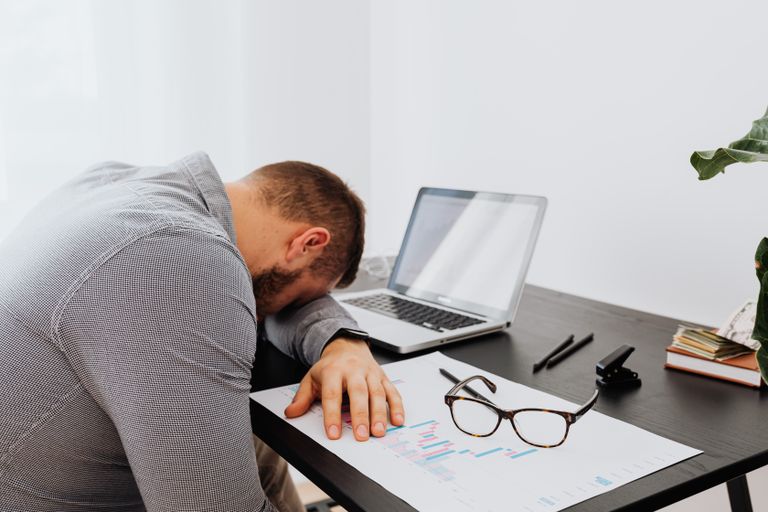 A man in a gray shirt sits tiredly at a desk with a laptop and documents, showing signs of workplace stress.