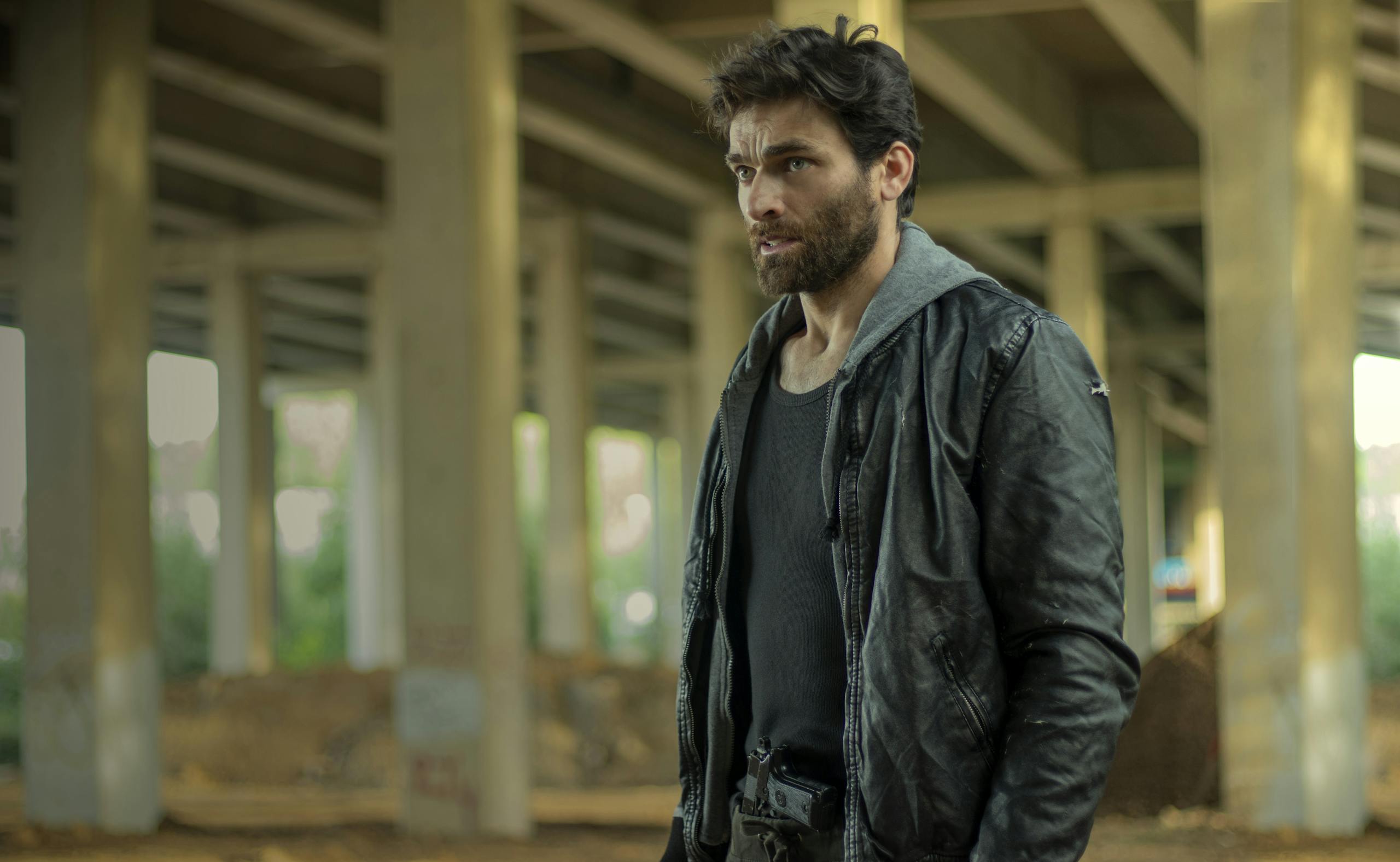 A man in a leather jacket stands under a bridge, embodying a gritty urban aesthetic.