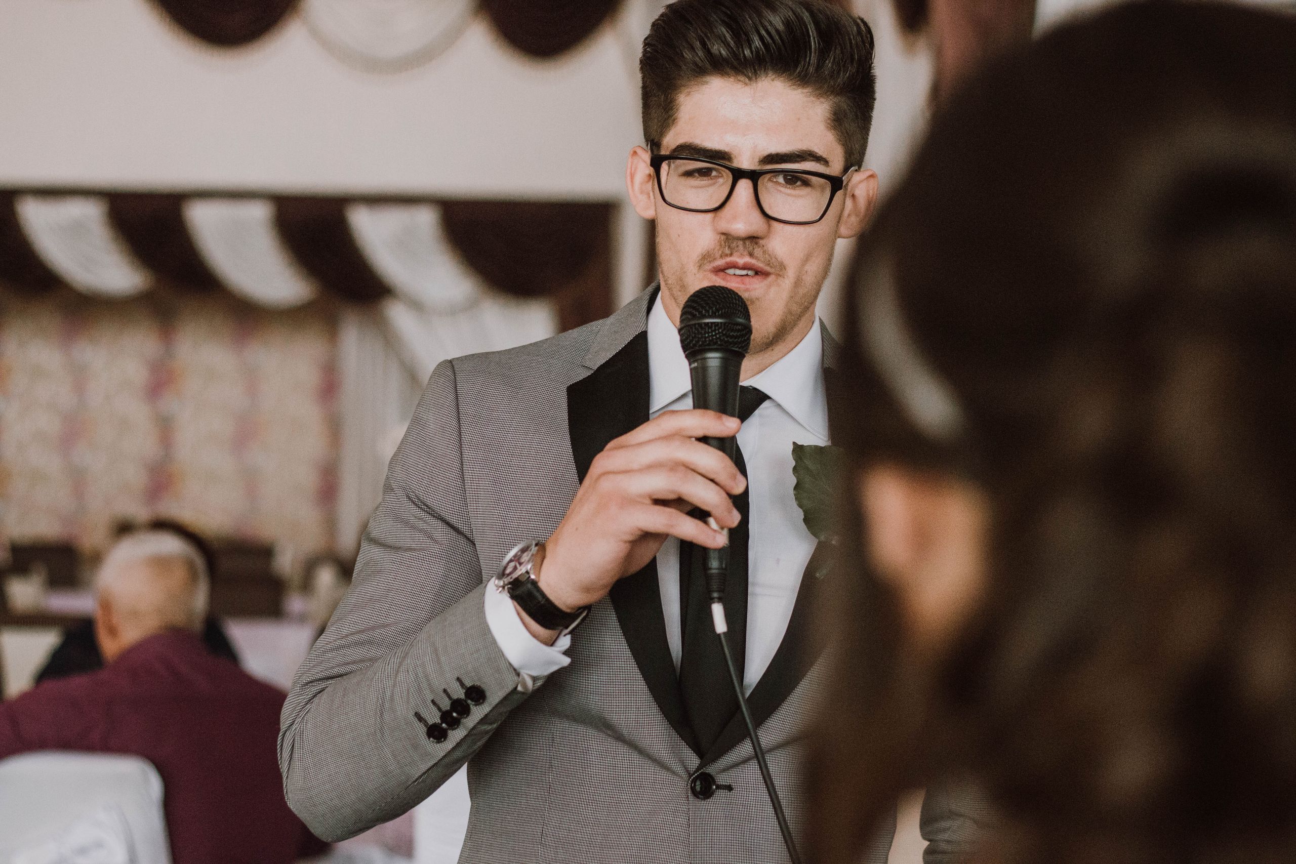 A man in a suit and glasses speaking into a microphone at a formal indoor event.