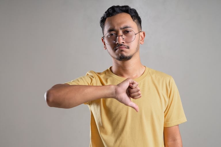 A man in glasses with a neutral expression giving a thumbs down gesture indoors.