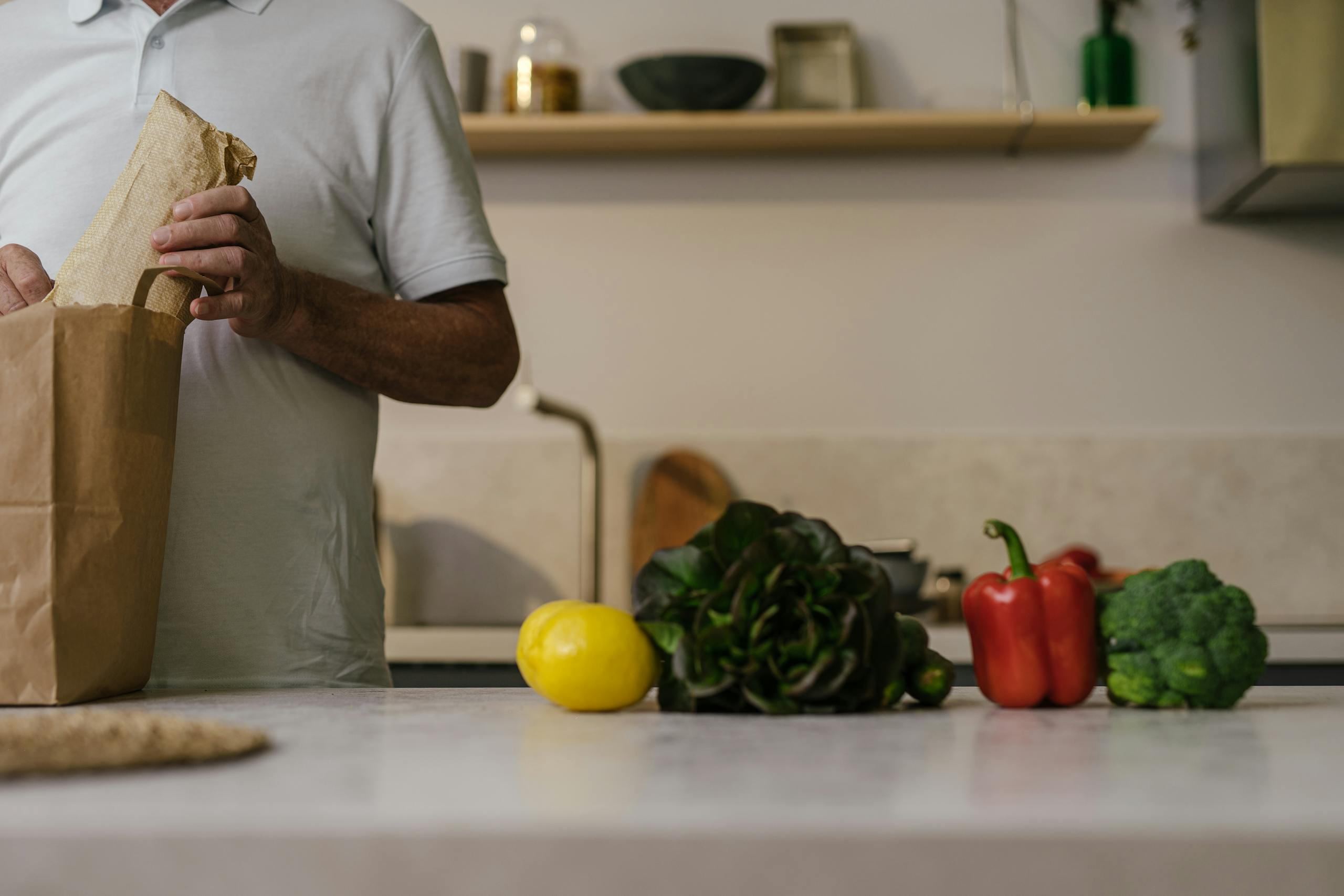 A man unpacks groceries from a paper bag on a kitchen counter with fresh vegetables.