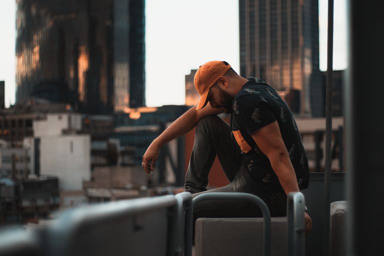 A man wearing a cap, sitting pensively on a rooftop with a city skyline at sunset.