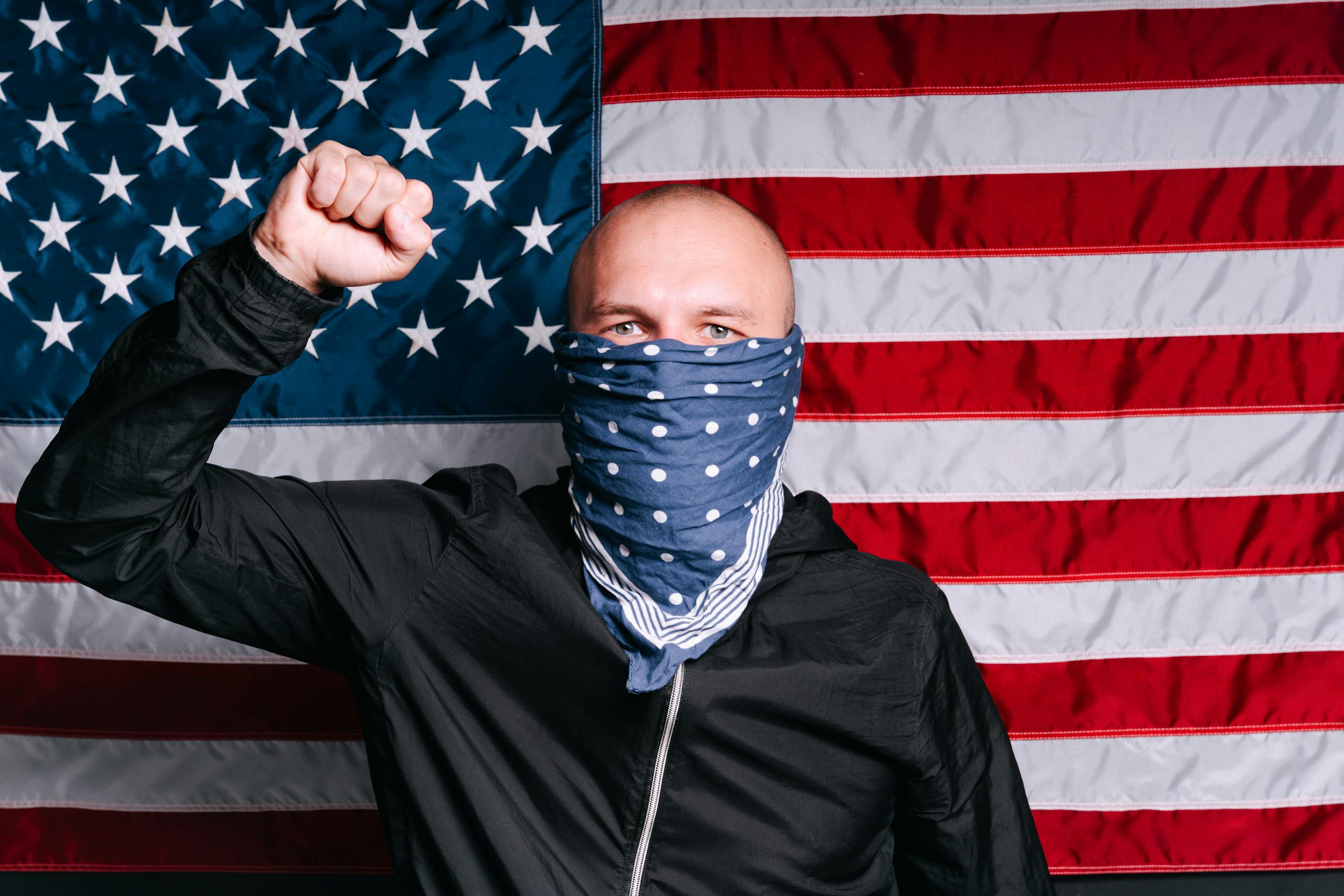A man wearing a mask stands with a raised fist in front of the American flag.