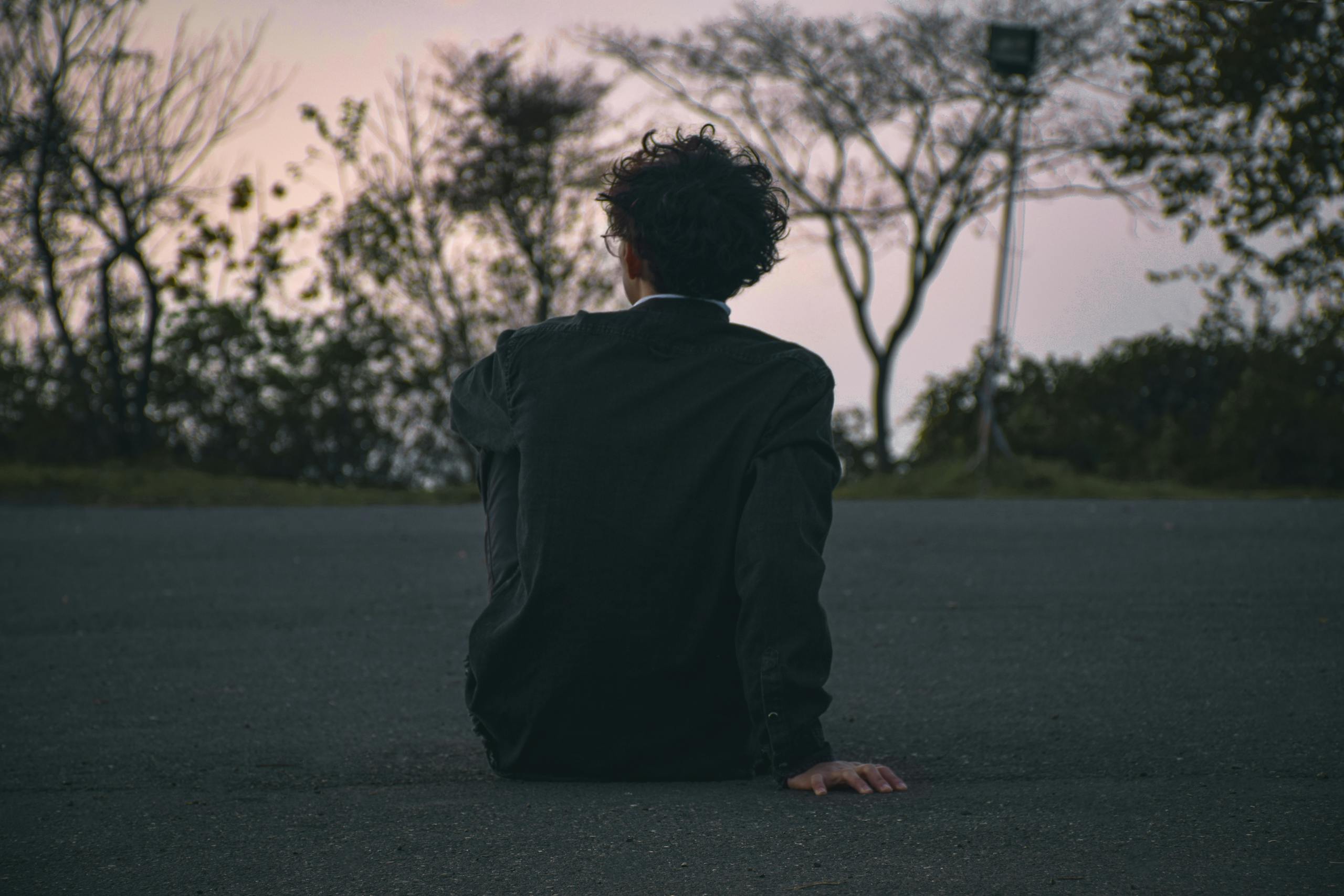 A man with curly hair sits on a road in solitude at dusk, offering a contemplative scene.