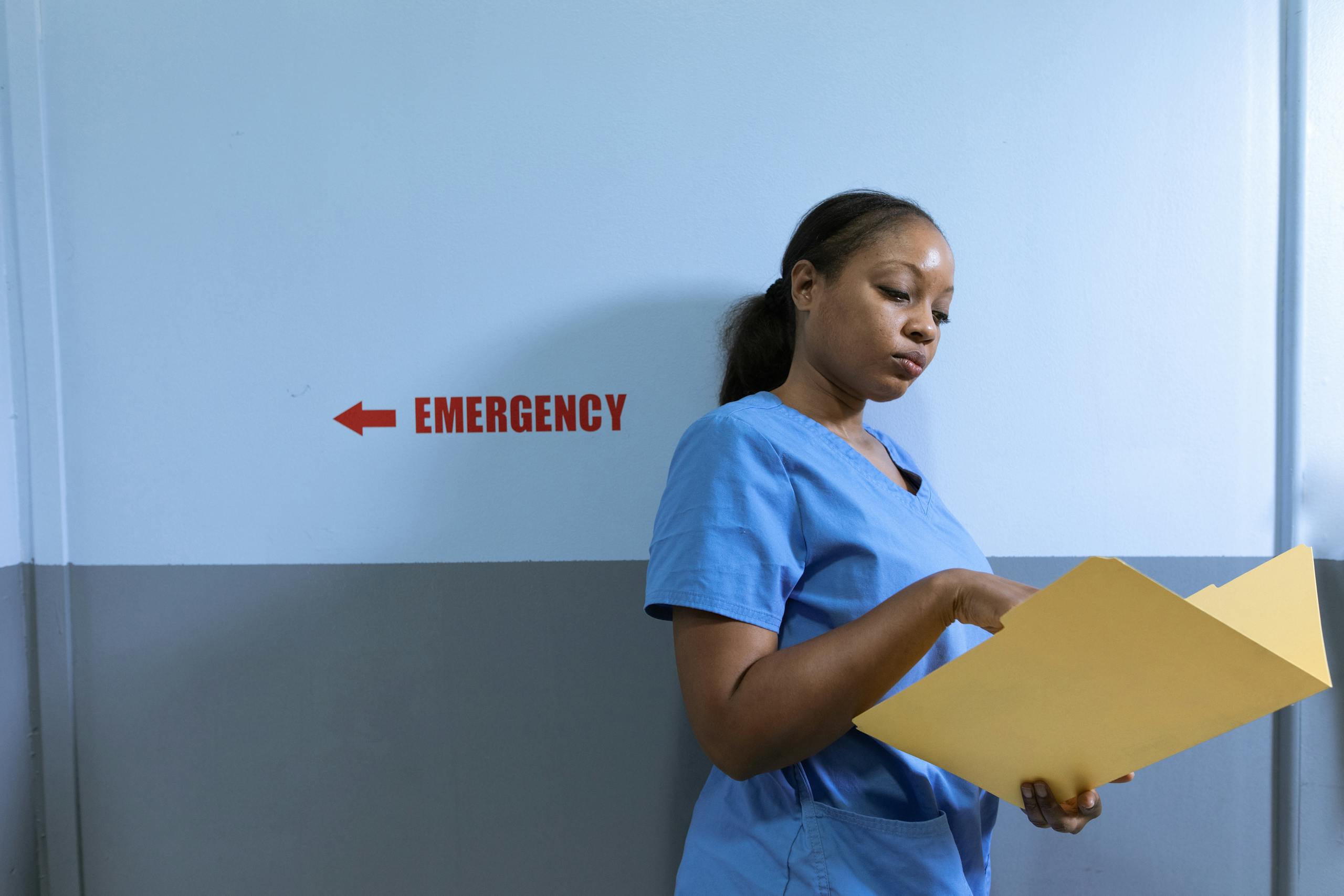 A nurse in blue scrubs reads a folder near an emergency room sign in a hospital.