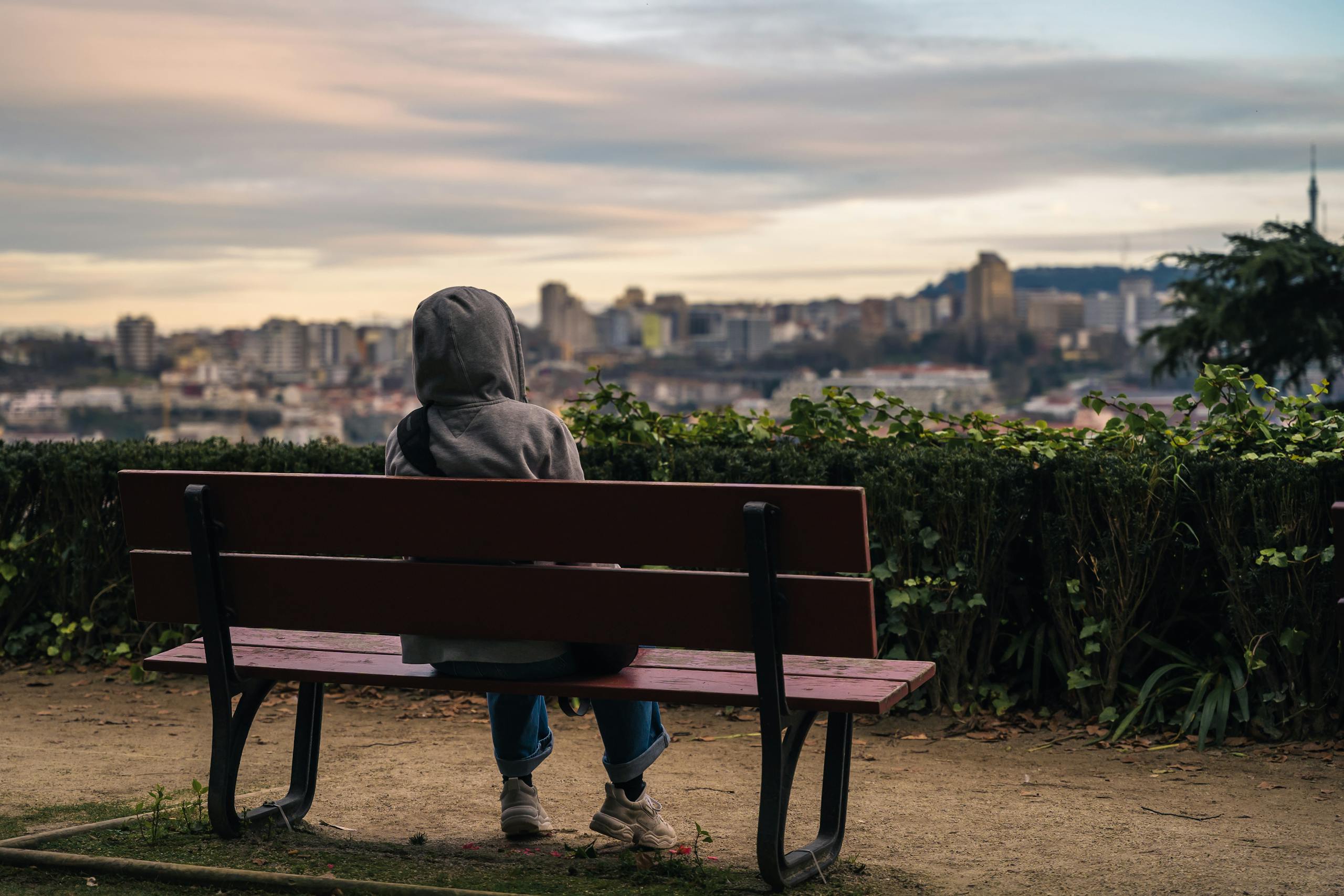 A person in a hoodie sits on a bench, gazing at the Porto cityscape during sunset.