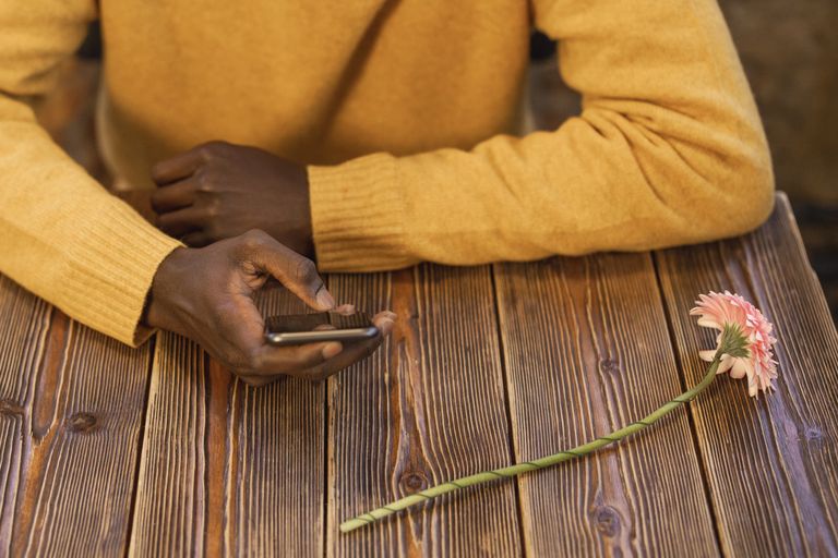 A person in a yellow sweater holds a smartphone on a rustic wooden table beside a single flower.