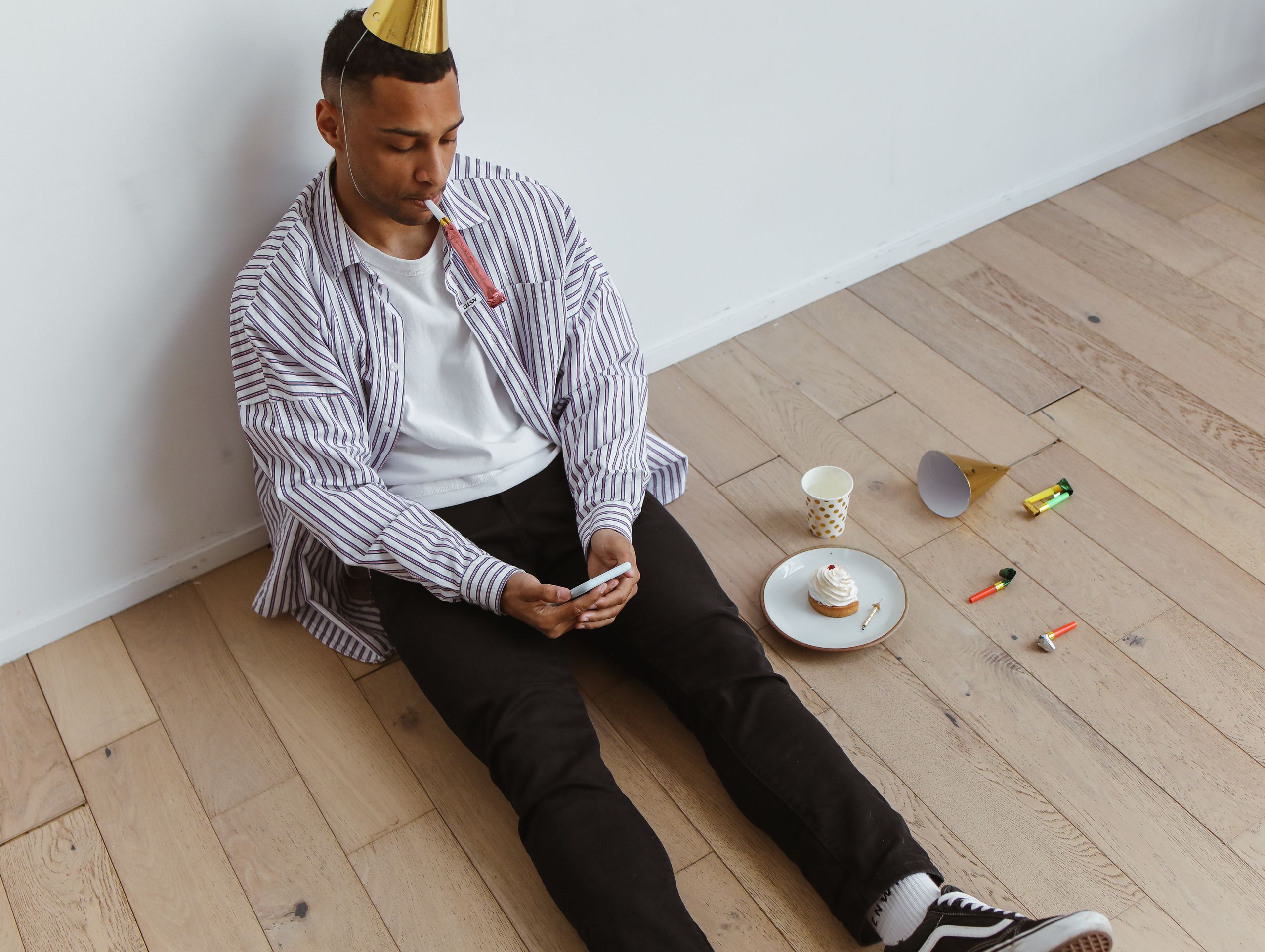 A person sits alone with a party hat, smartphone, and cupcake, depicting solitude in celebration.
