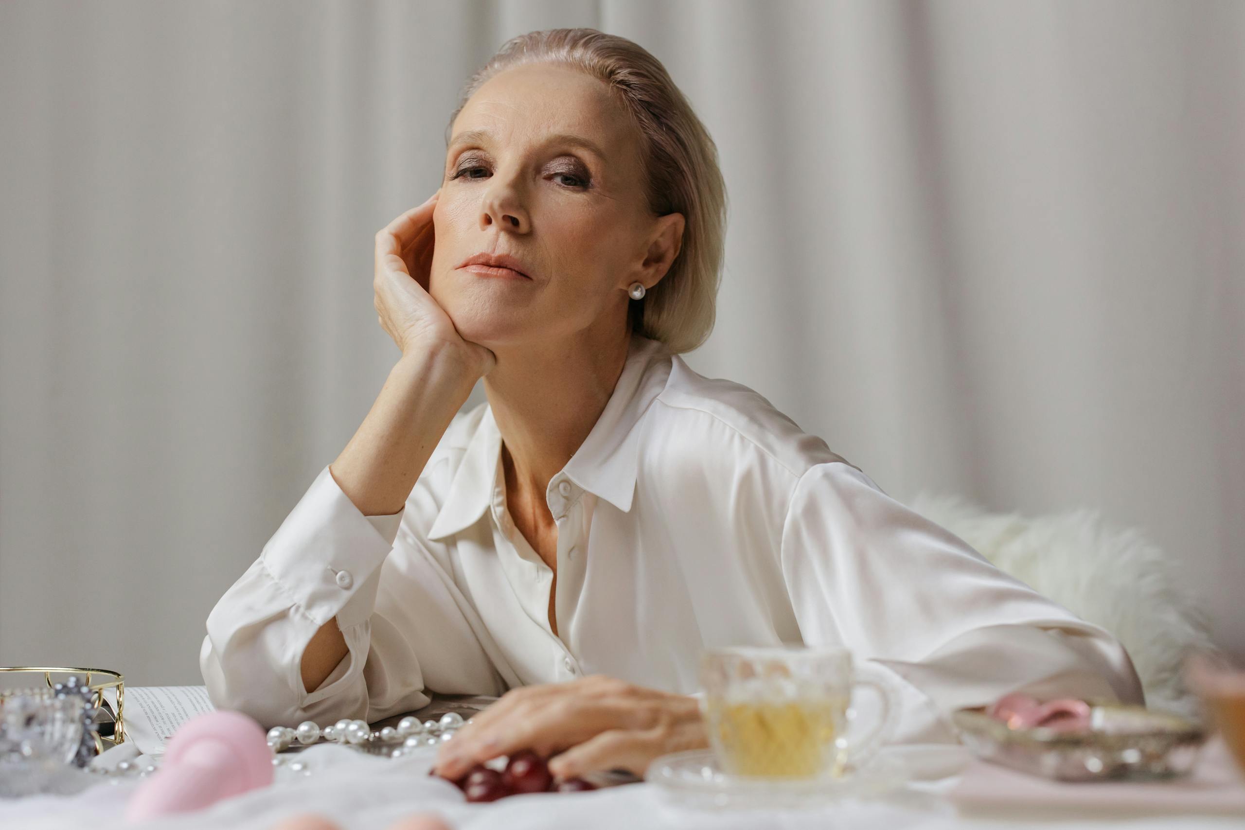 A poised senior woman in a white shirt enjoying tea, surrounded by elegant pearls.