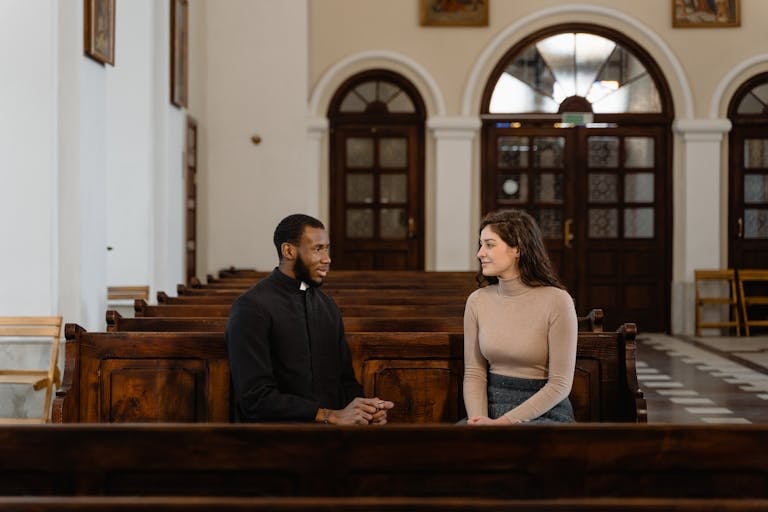 A priest and a woman in conversation sitting in church pews, creating a serene and introspective scene.