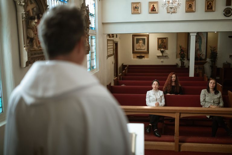 A priest leads a church service as three congregants listen attentively in the pews.