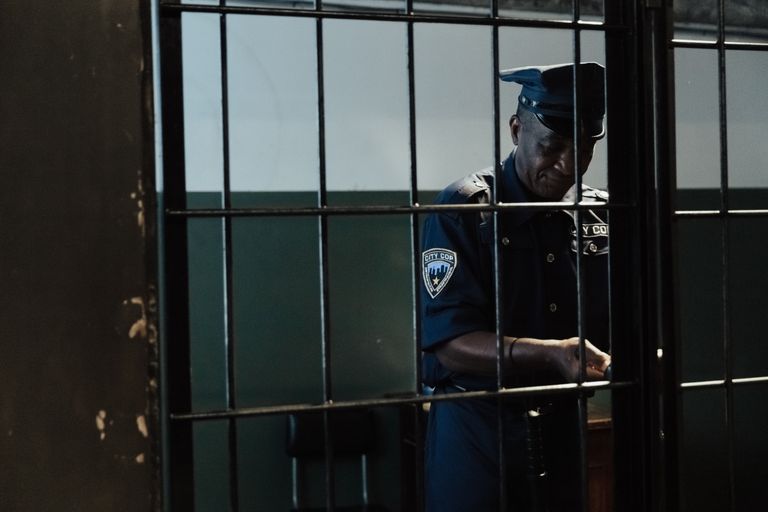 A security officer in uniform standing behind bars in a dimly lit room, evoking themes of law enforcement and confinement.
