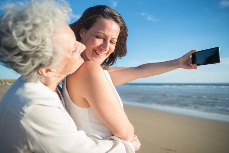 A senior mother and adult daughter taking a joyful selfie on a sunny beach in Portugal.
