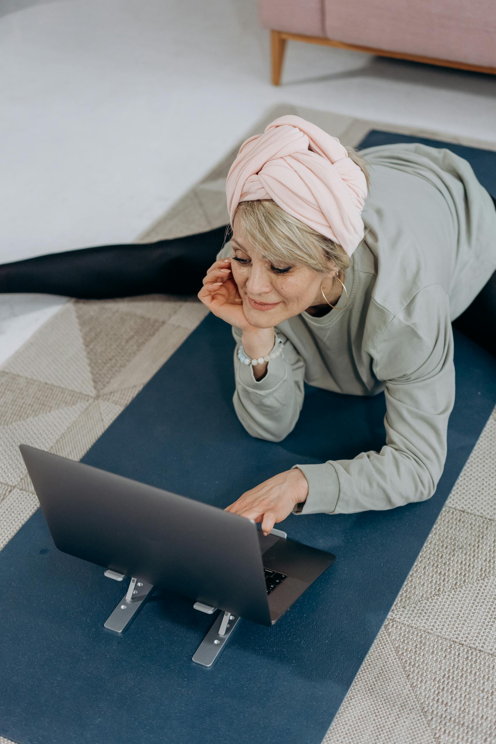 A senior woman with a headwrap using a laptop on a yoga mat for online fitness exercises.