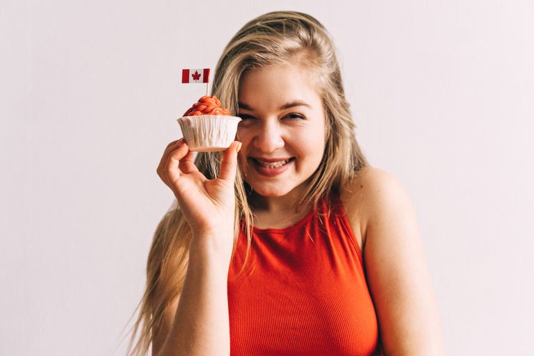 A smiling woman in a red top holding a cupcake with the Canadian flag on a white background.