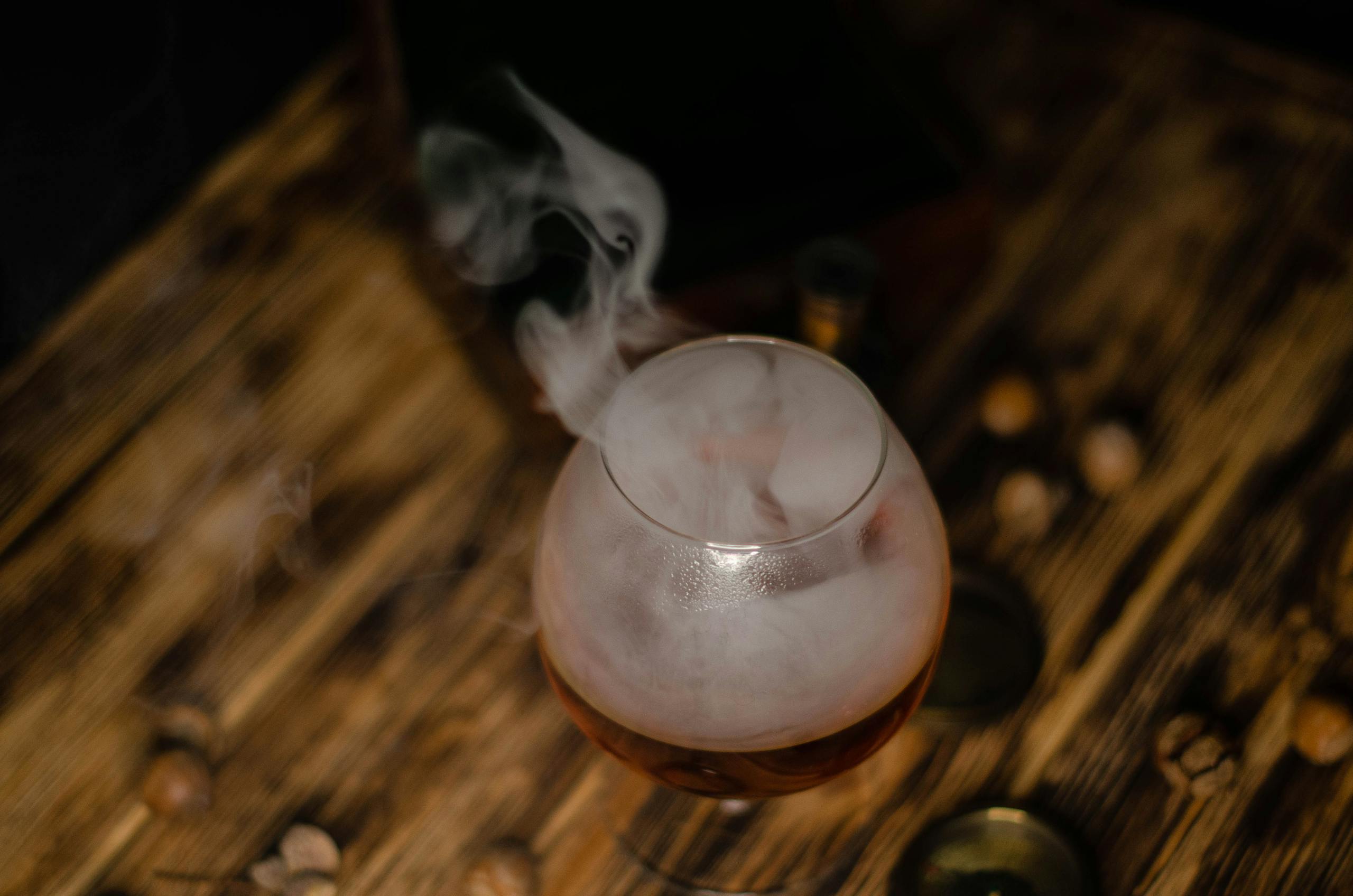 A smoky cocktail served in a glass on a rustic wooden table, surrounded by nuts and smoke.