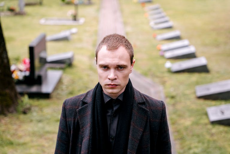 A somber man dressed in black standing in a cemetery, reflecting on loss and grief.