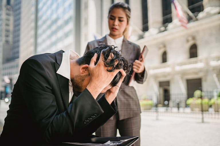 A stressed businessman holds his head in despair as his female colleague stands by him outdoors.