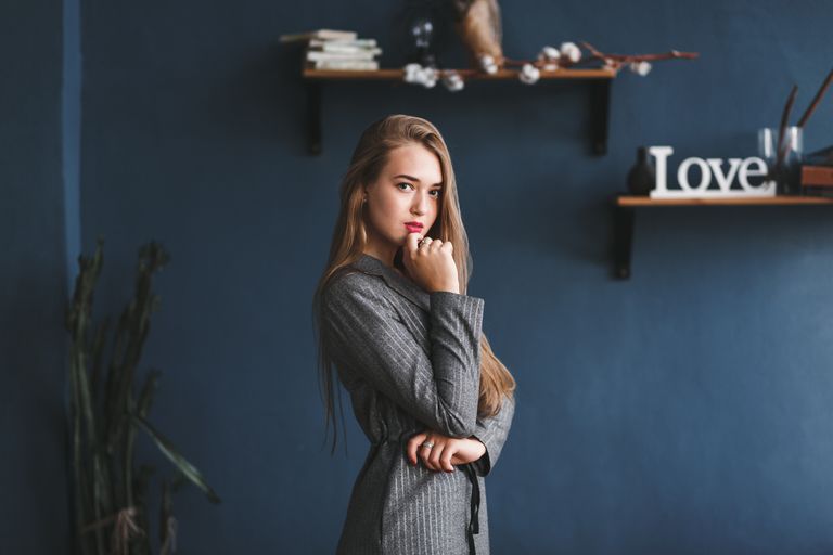 A stylish caucasian woman poses thoughtfully indoors, wearing a blazer against a dark artistic background.