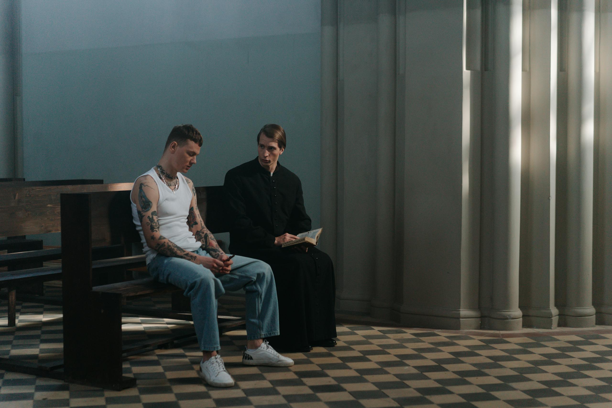 A tattooed man seeks counsel from a priest in a serene church interior, highlighting themes of faith and reflection.