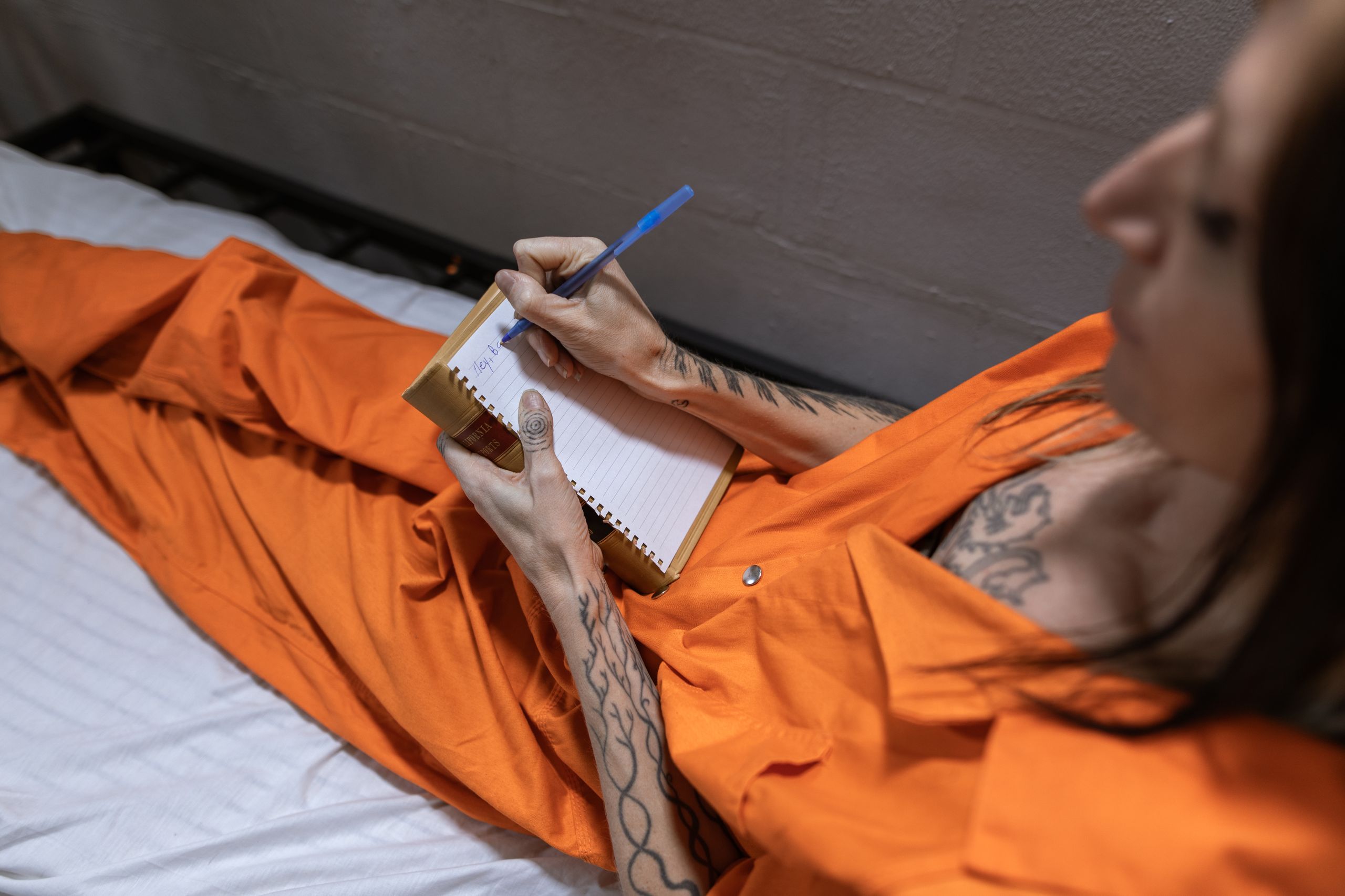 A tattooed woman in an orange jumpsuit writes in a notebook, symbolizing introspection and rehabilitation.
