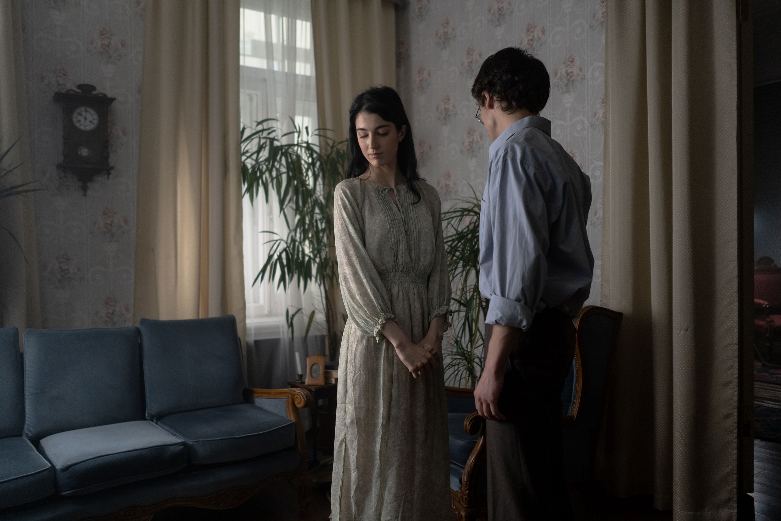 A thoughtful couple in vintage attire stands indoors surrounded by retro decor.