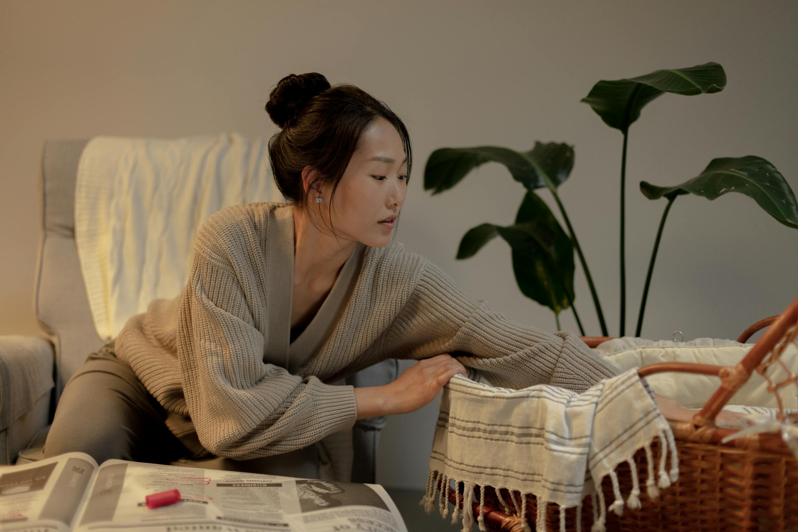 A thoughtful woman leans over a bassinet in a cozy home interior setting.