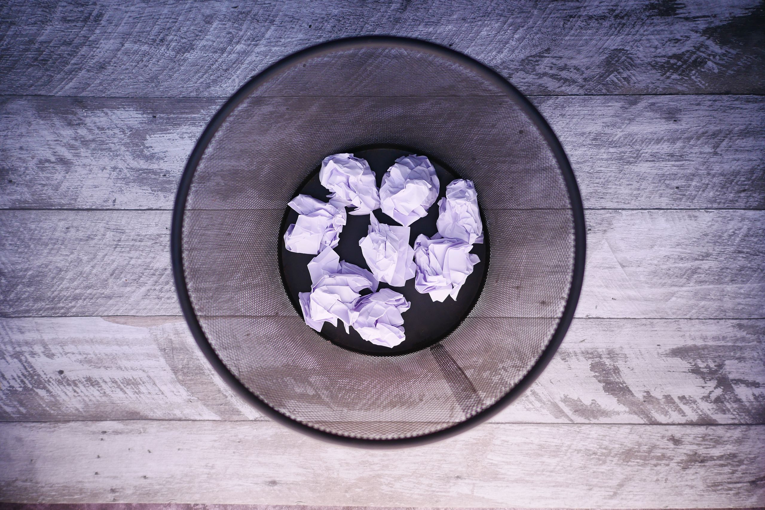 A top-down view of crumpled papers inside a black trash can on wooden flooring.
