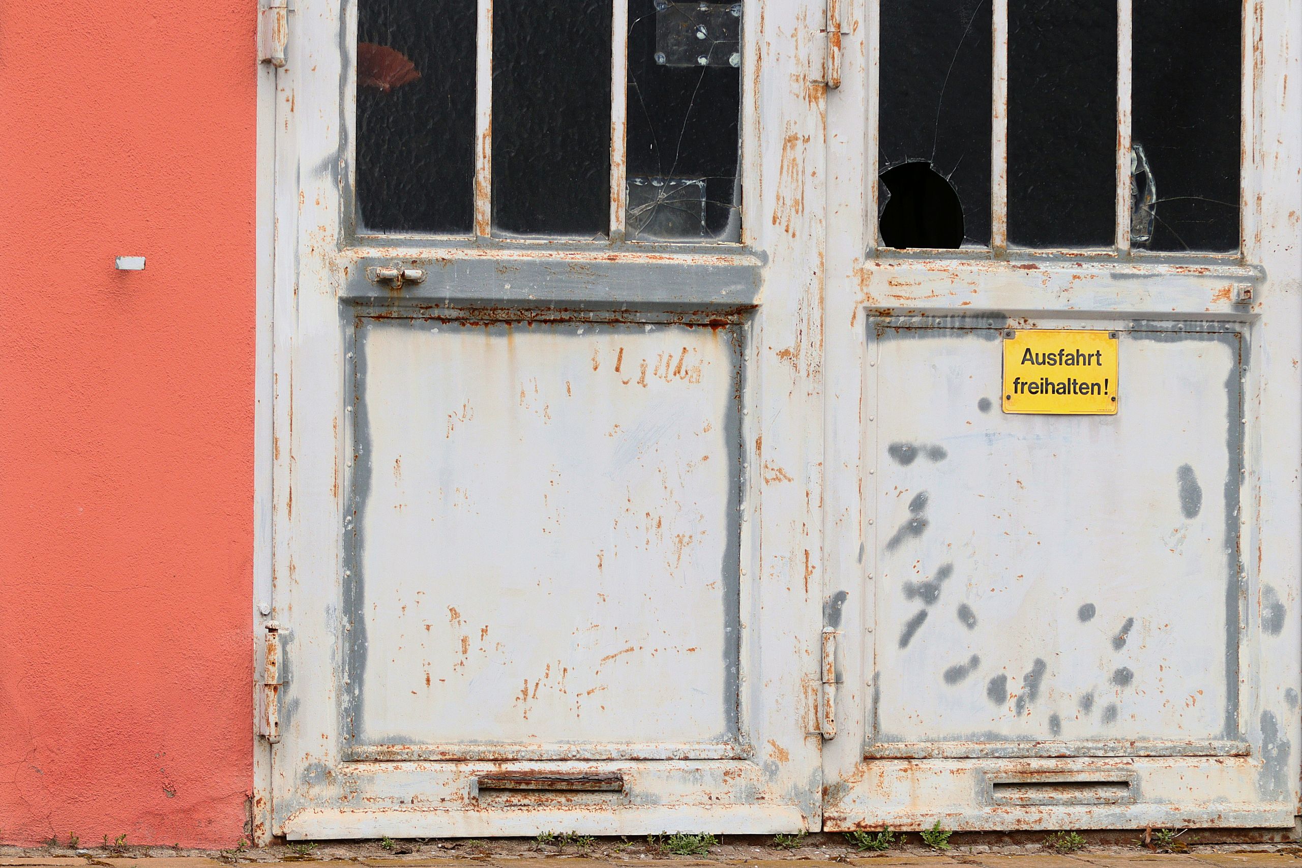 A weathered, rusty door with broken glass panes and a yellow warning sign in an urban setting.