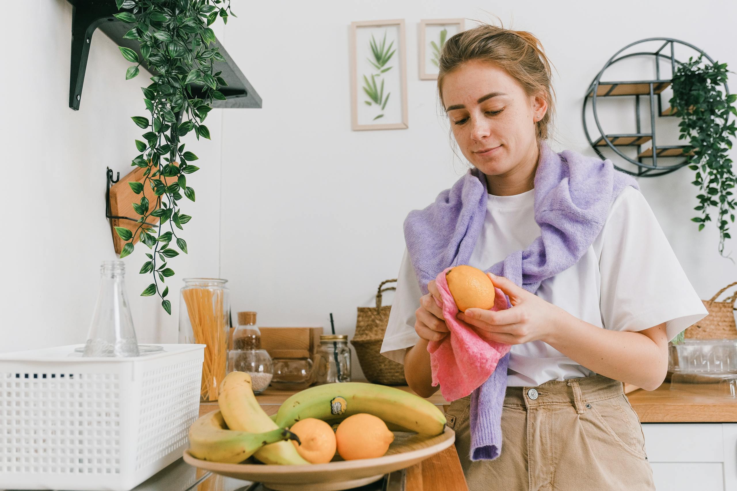 A woman cleaning fresh oranges in a modern kitchen, surrounded by bananas and plants.