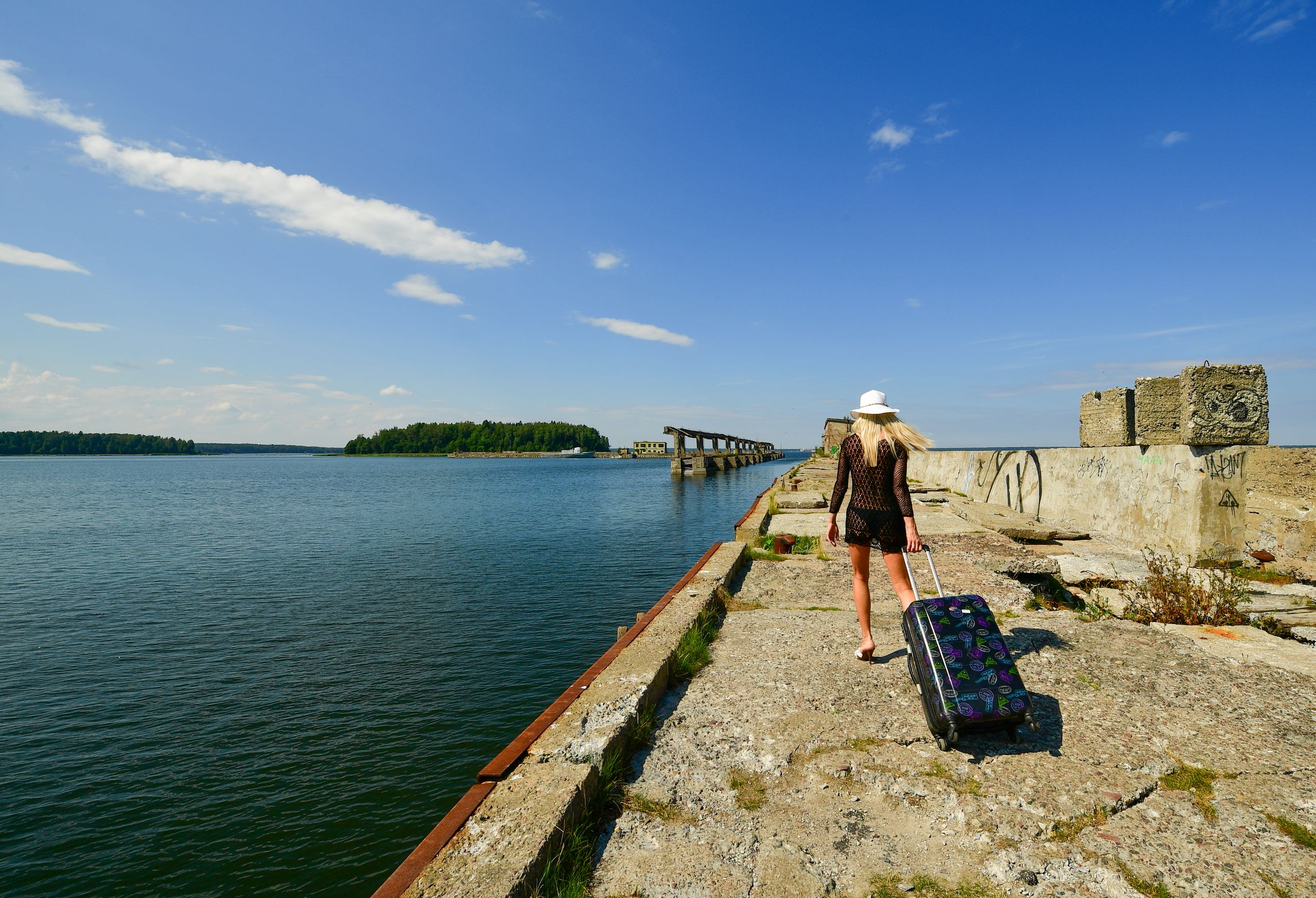 A woman in a black dress and white hat walks on an abandoned pier in Hara, Estonia, under a sunny blue sky.