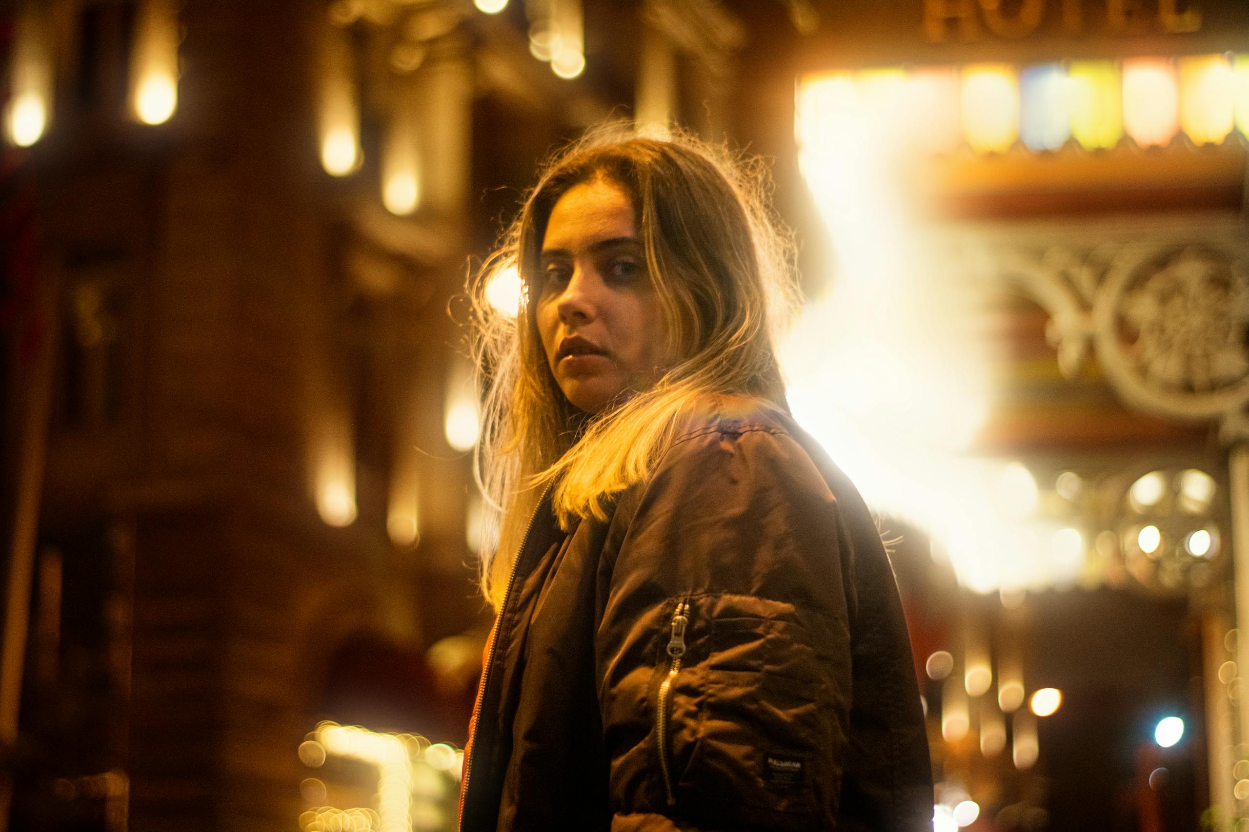 A woman in a black jacket stands on a bustling, illuminated city street at night, looking back.
