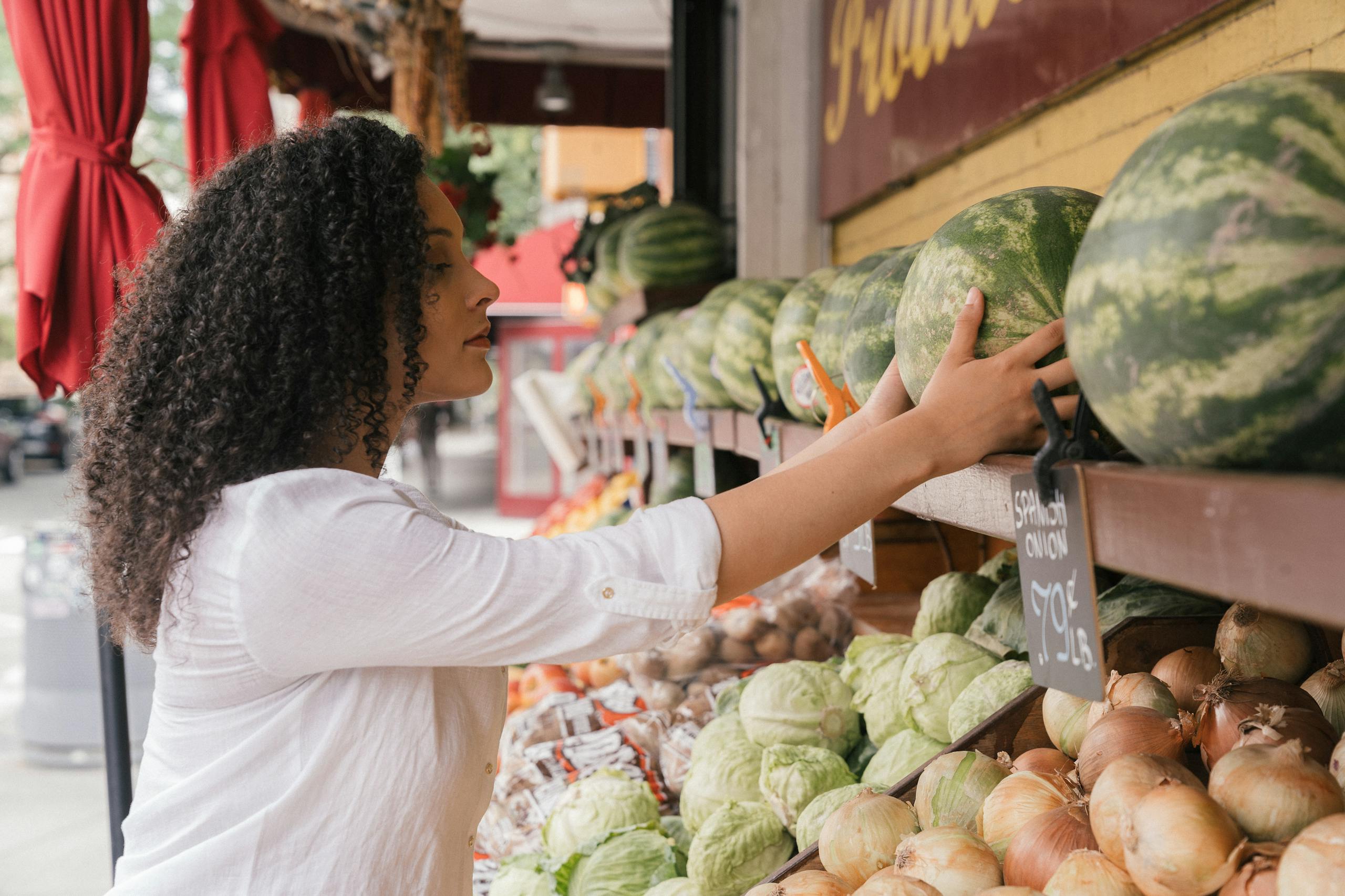 A woman in a white top choosing fresh watermelons at an outdoor market stall.