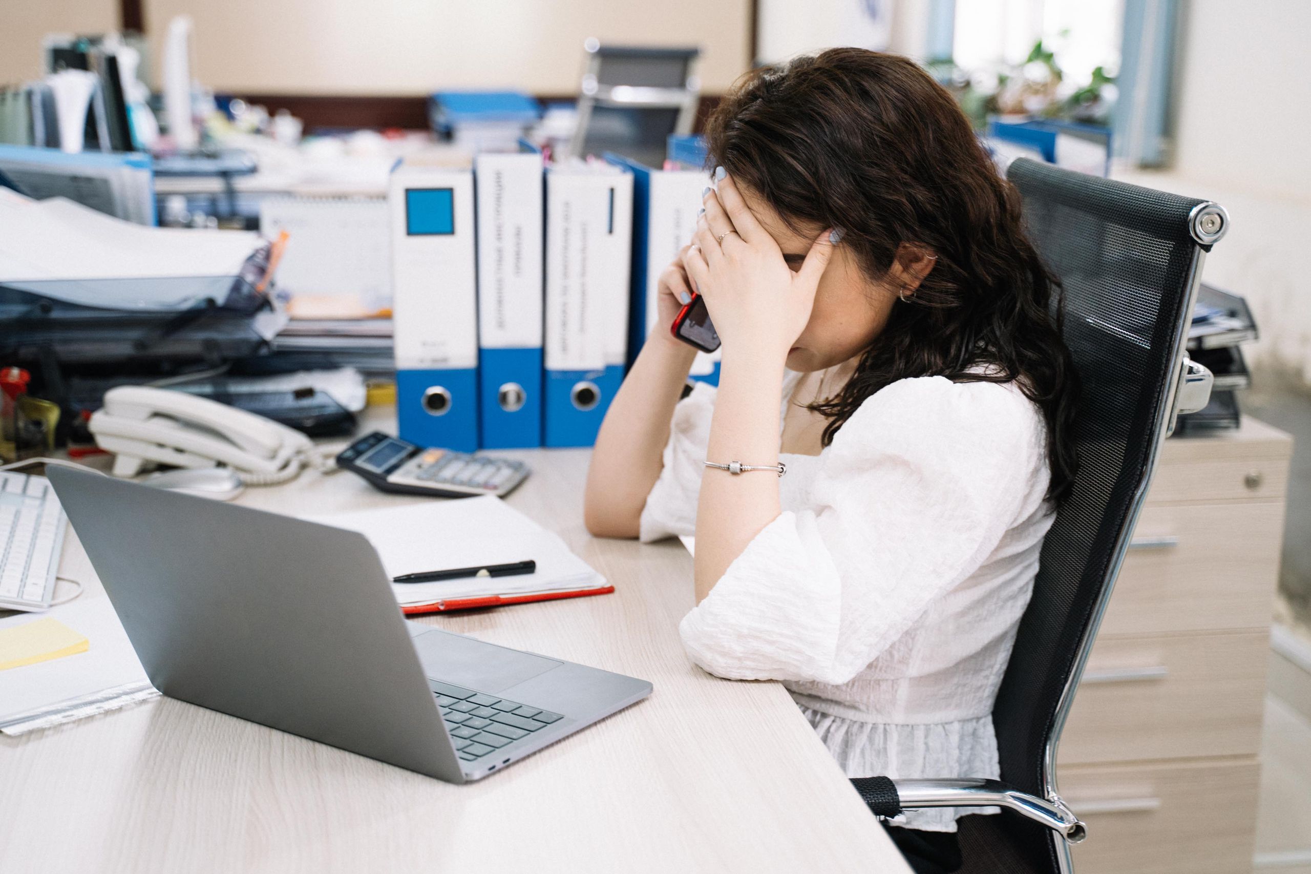 A woman in an office sitting at a desk, visibly stressed, using a phone. Workplace stress concept.