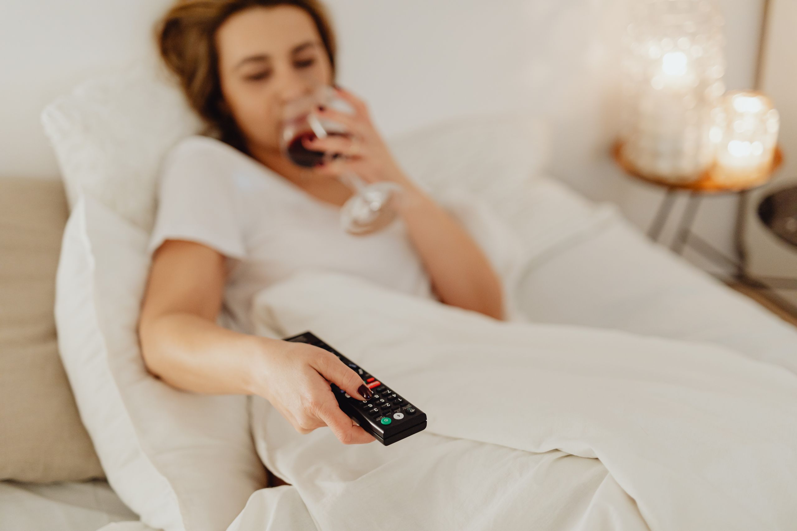 A woman in bed holding a glass of wine and a remote control, enjoying a cozy evening.