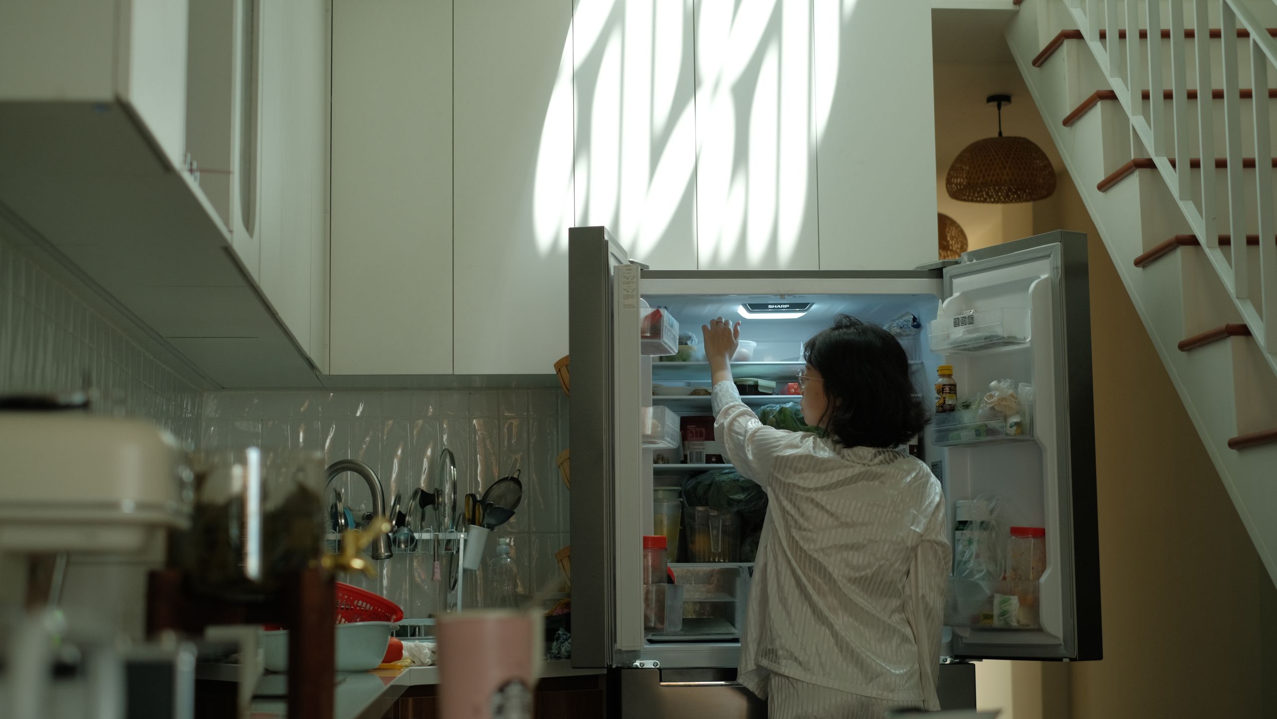 A woman in pajamas checks the refrigerator in a warmly lit cozy kitchen.