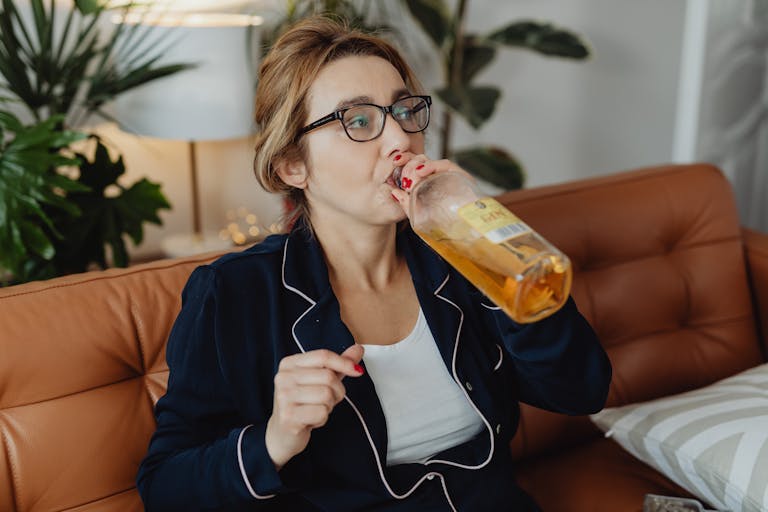 A woman in pajamas sits on a couch, drinking from a glass bottle indoors.