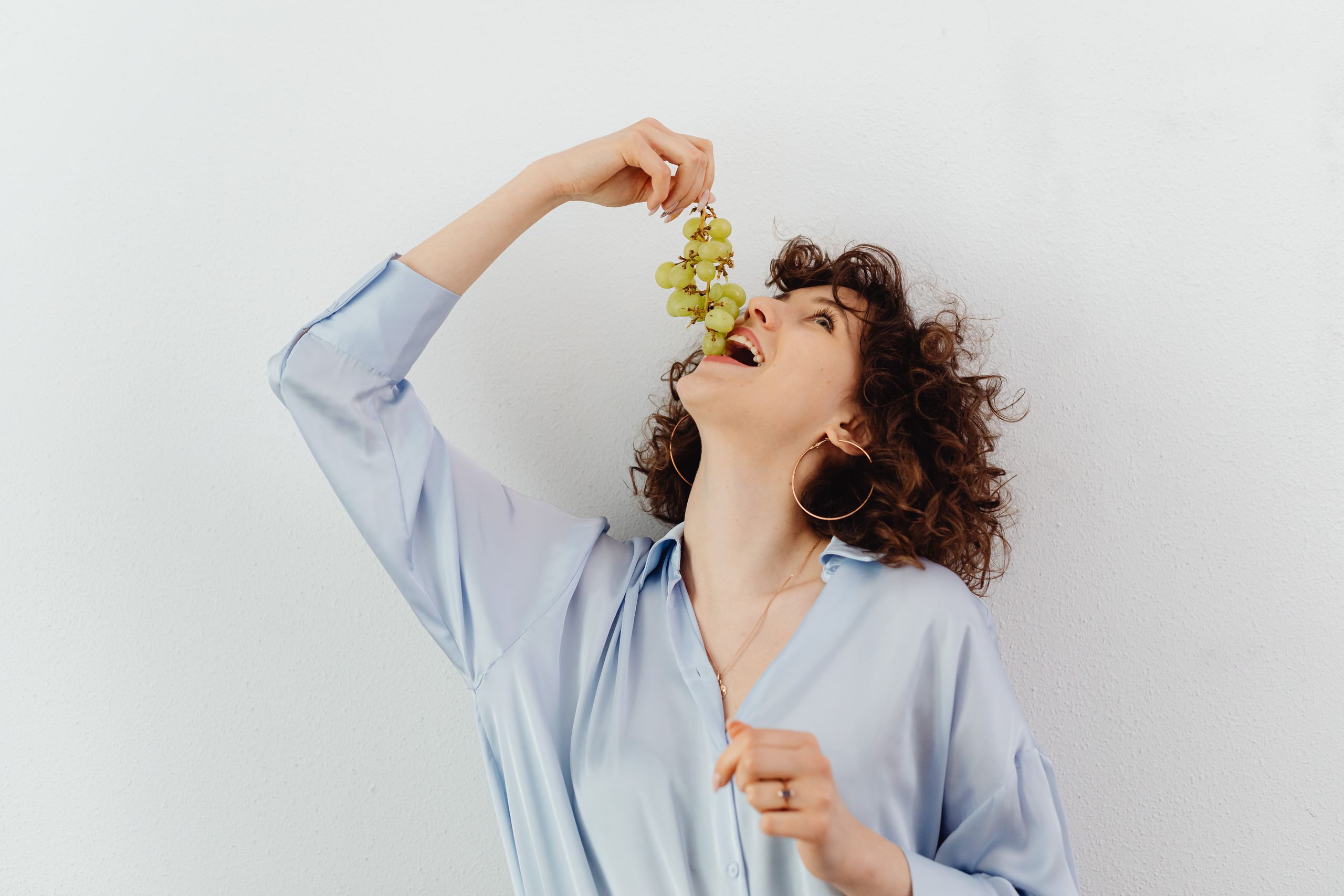 A woman joyfully eating green grapes, wearing a blue blouse, indoors.