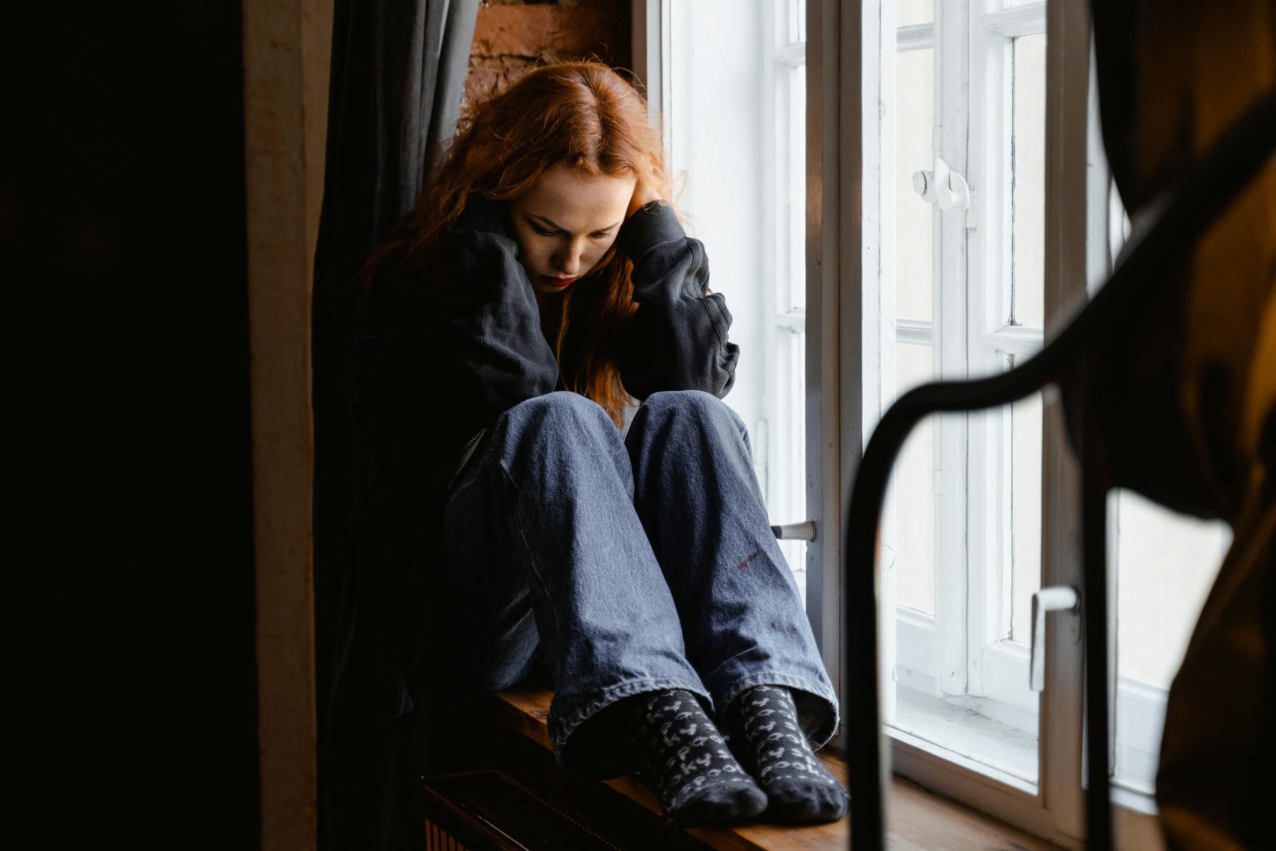 A woman sits pensively by a window, expressing feelings of solitude and contemplation.